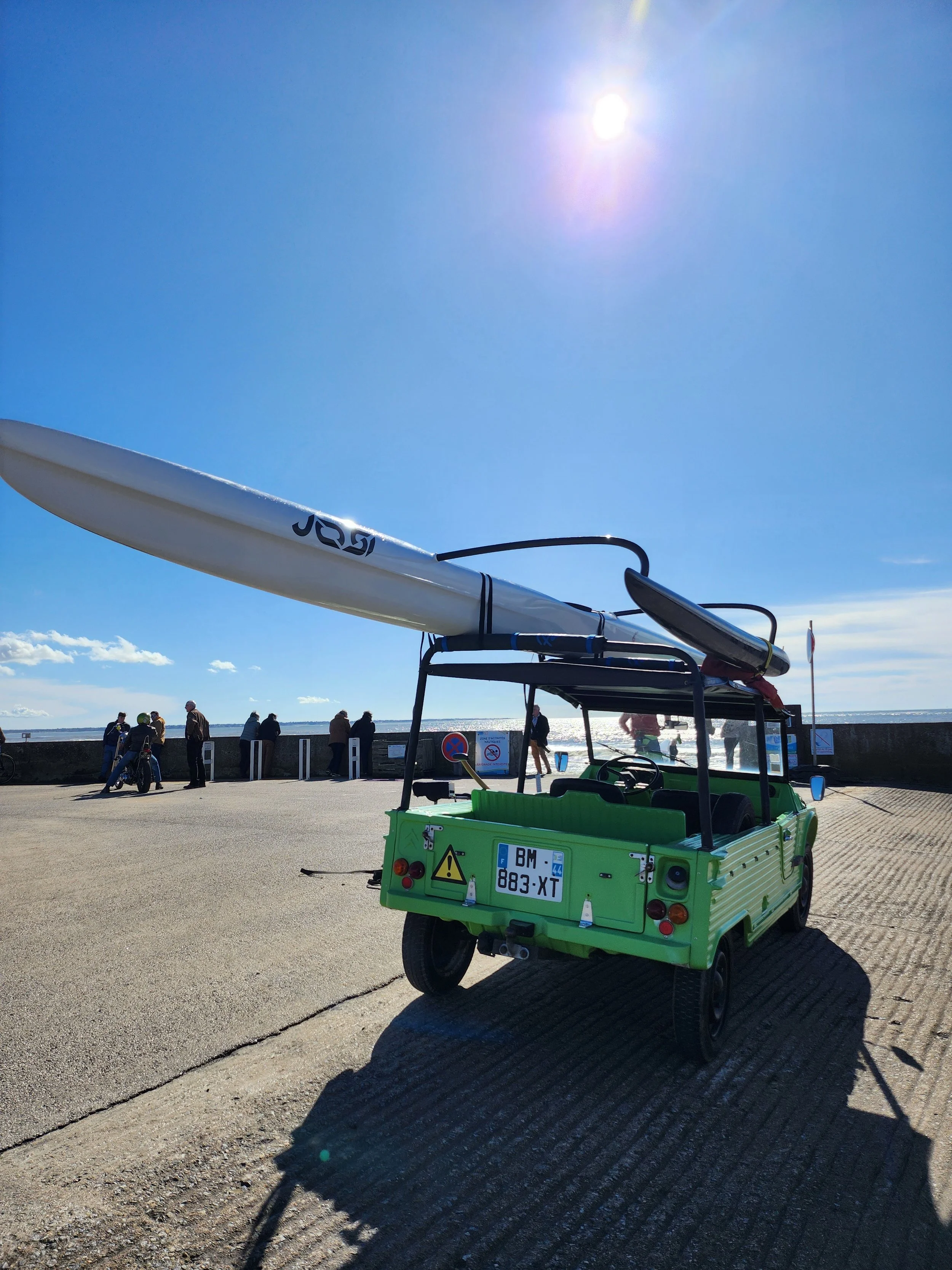 Voiture verte avec un kayak blanc attaché sur le toit, stationnée près de la mer par une journée ensoleillée, avec des personnes en arrière-plan regardant la mer ou se promenant sur la promenade.