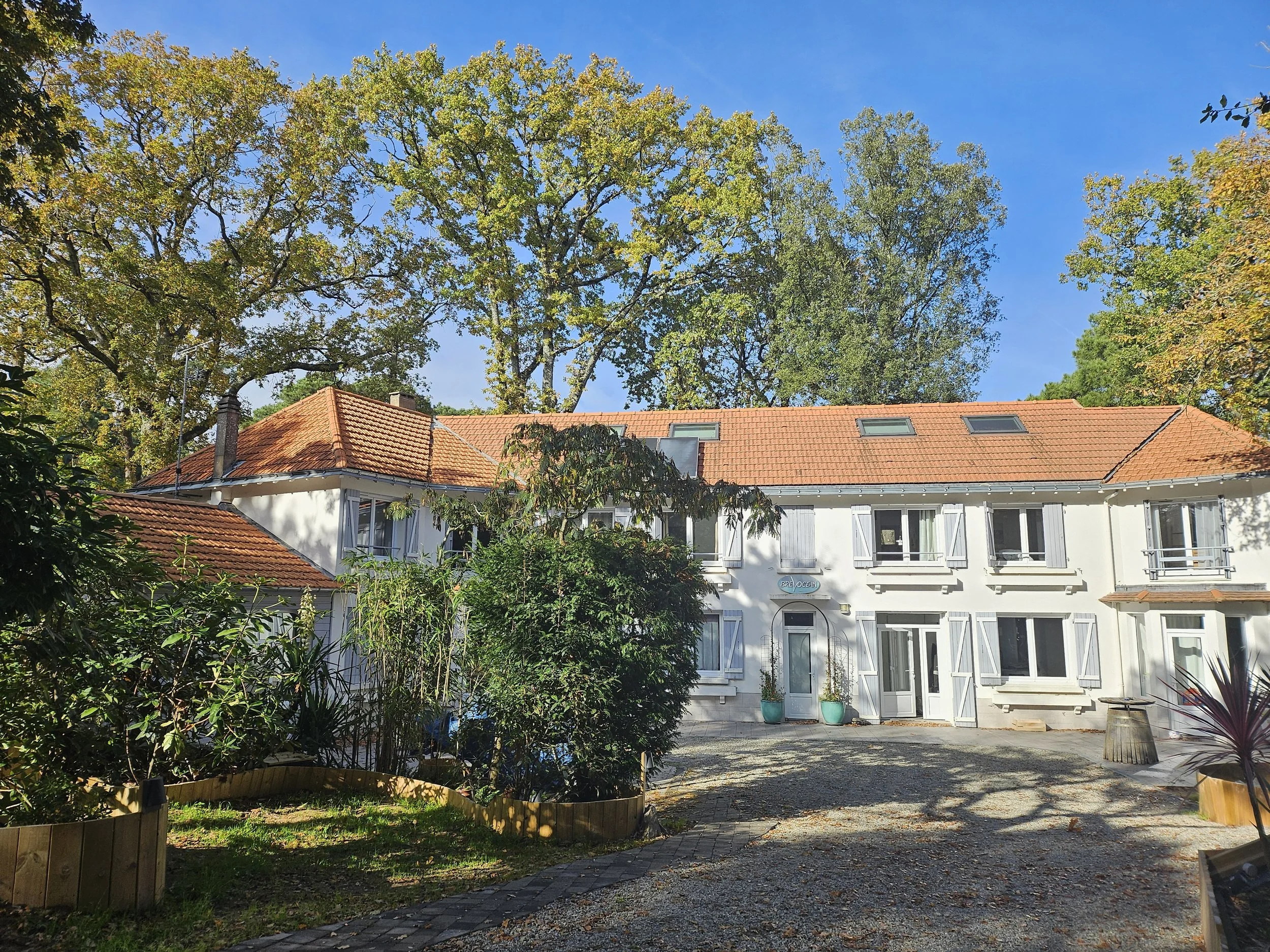 Maison blanche à deux étages avec volets gris, toiture en tuiles rouges, entourée d'arbres et de végétation, sous un ciel bleu.