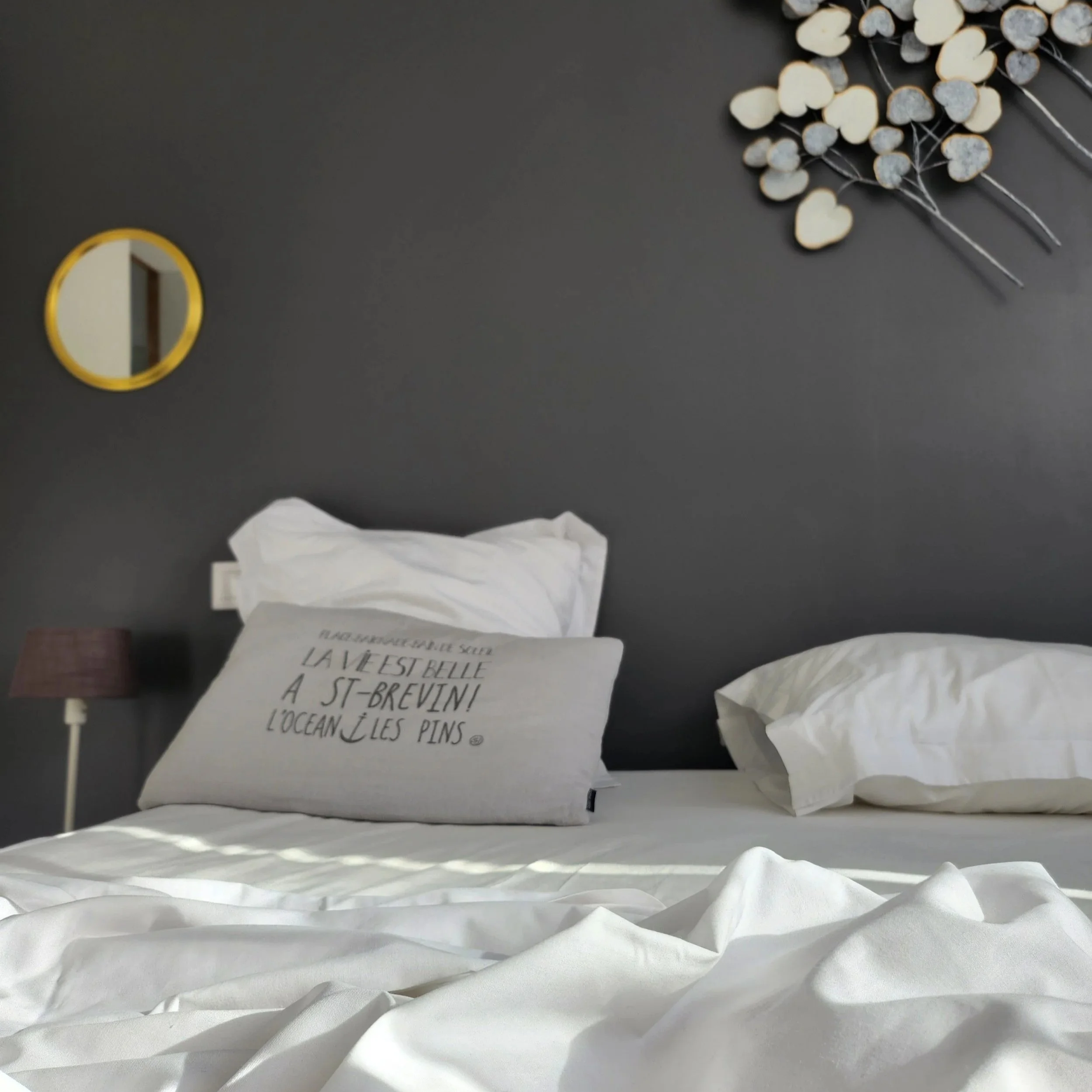 Un lit avec deux oreillers blancs sur un drap blanc, un mur gris foncé en arrière-plan, une petite lampe de table mauve à gauche, un miroir rond avec cadre jaune à gauche, et une décoration murale en forme de coeurs blancs et gris en haut à droite.