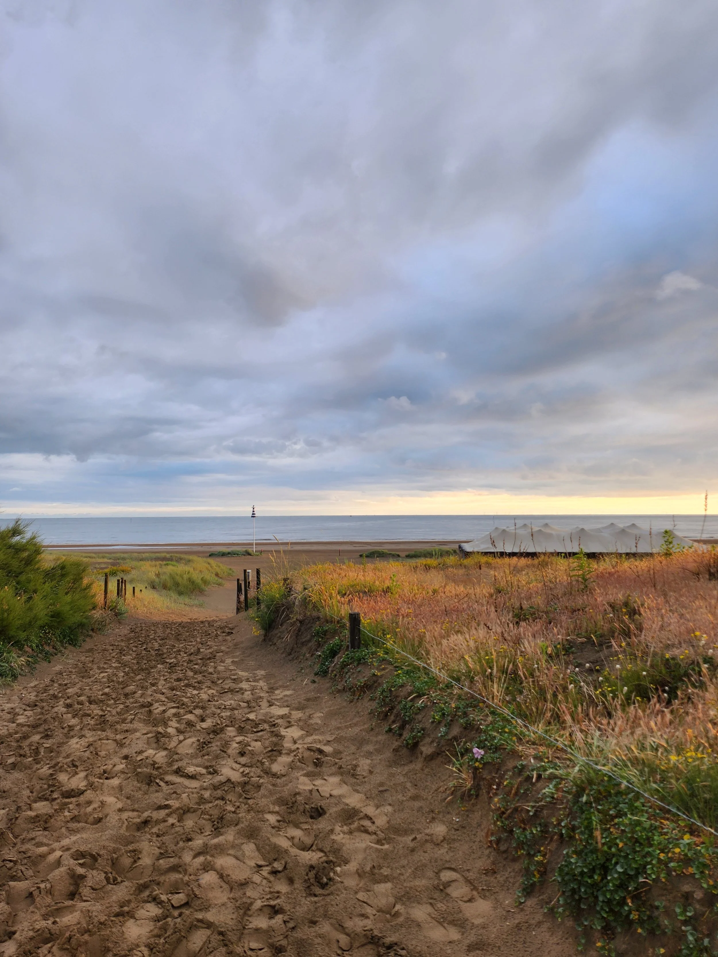 Chemin de sable menant à la plage verte de Saint-Brevin-l'océan avec dans les dunes des herbes appelés oyats, un ciel nuageux et la mer côte atlantique à l'horizon.