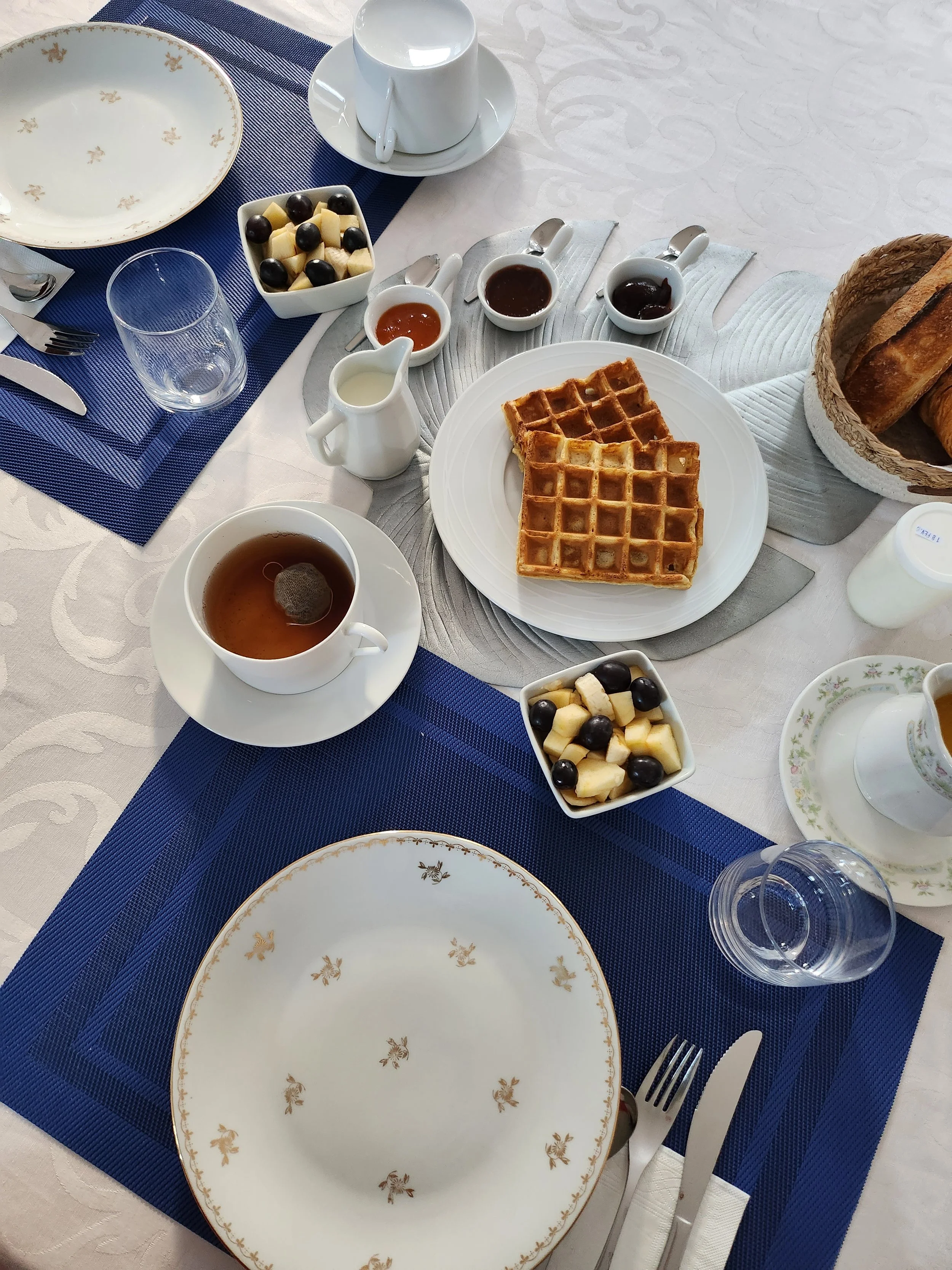 Table de petit déjeuner avec des gaufres maison, du beurre, du pain, des fruits, des tasses de thé et de café, produits locaux et des assiettes décorées de motifs dorés.