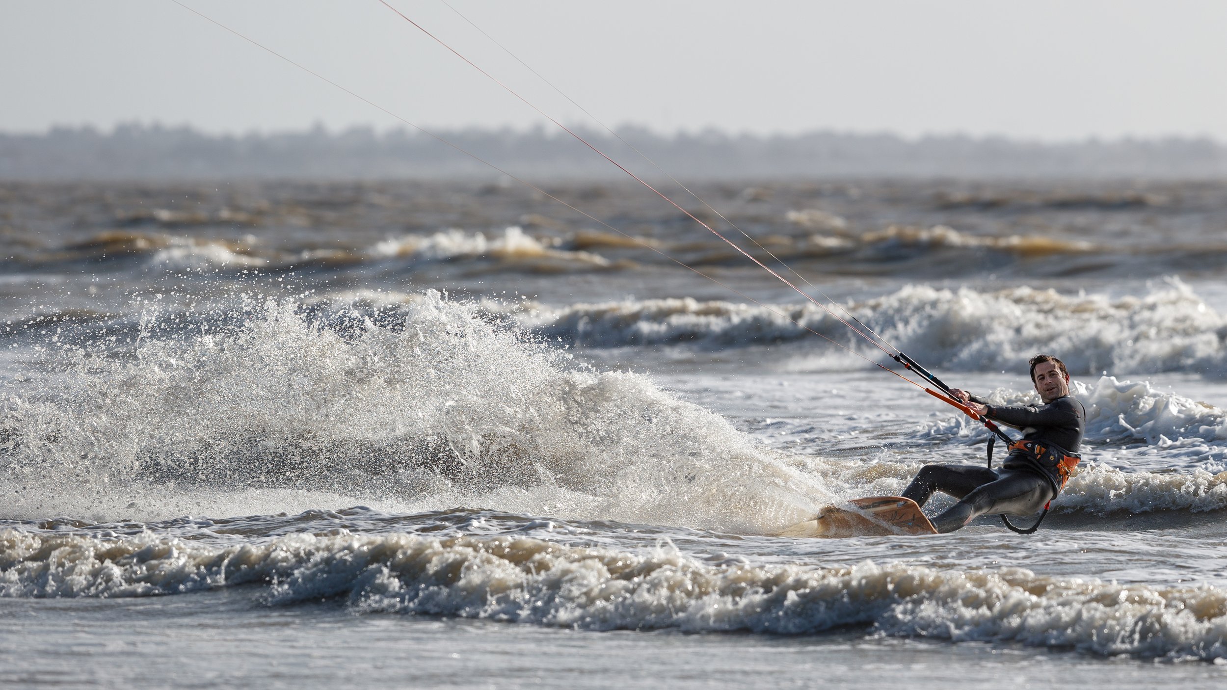 Un homme en combinaison de plongée pratique la planche à voile dans la mer, tirant sur la voile avec la voile flottant dans le vent, créant une éclaboussure d'eau.