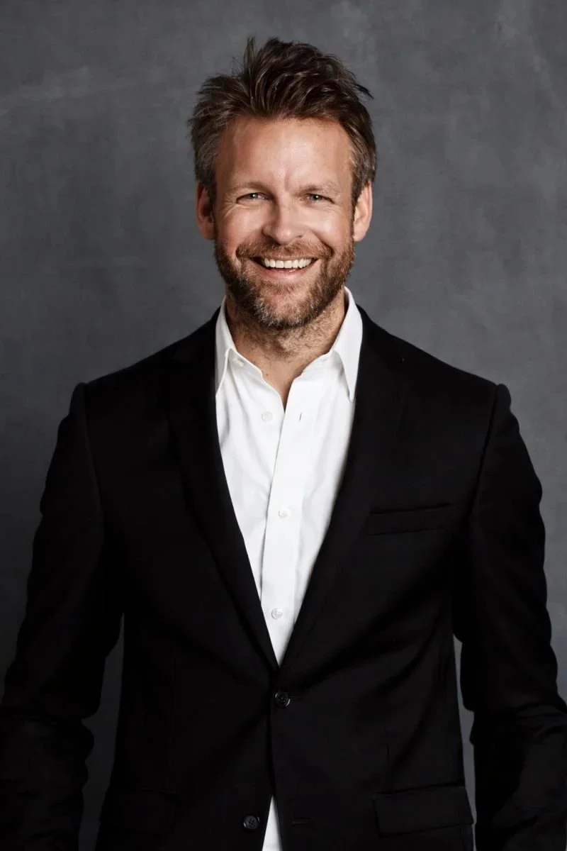 En mann med skjegg og brunt, litt bustete hår smiler mot kamera, iført sort dress og hvit skjorte, mot en grå bakgrunn.