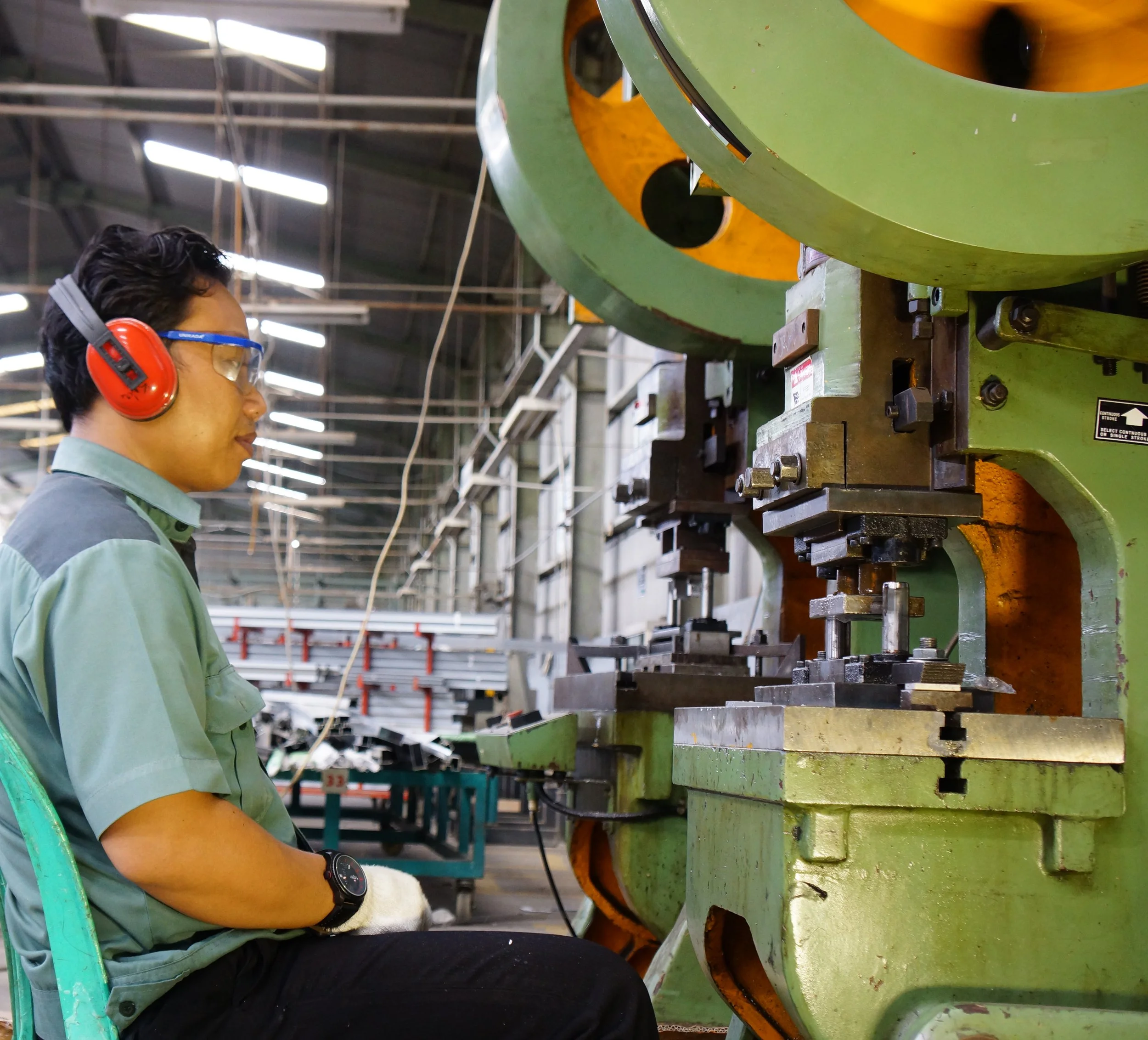 A worker wearing safety glasses and red ear protection operating a large industrial machine in a manufacturing factory.