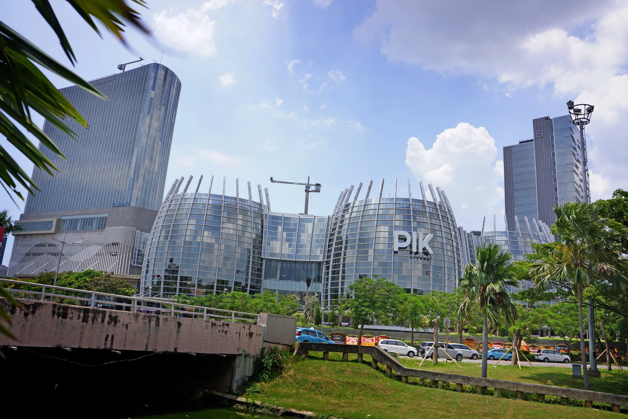 Modern cityscape featuring the PIK Avenue shopping mall with its distinctive curved glass exterior, surrounded by lush greenery and tall skyscrapers under a partly cloudy sky.