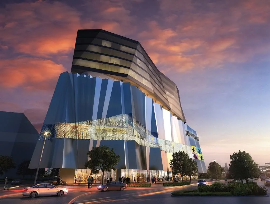 A modern multi-story building with a unique, angular facade and glass exterior, illuminated at dusk with trees and cars in the foreground.