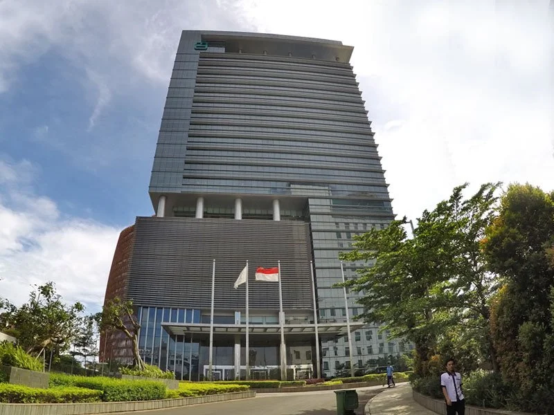 Tall modern office building with multiple glass windows, surrounded by trees and a pathway, with flags in front