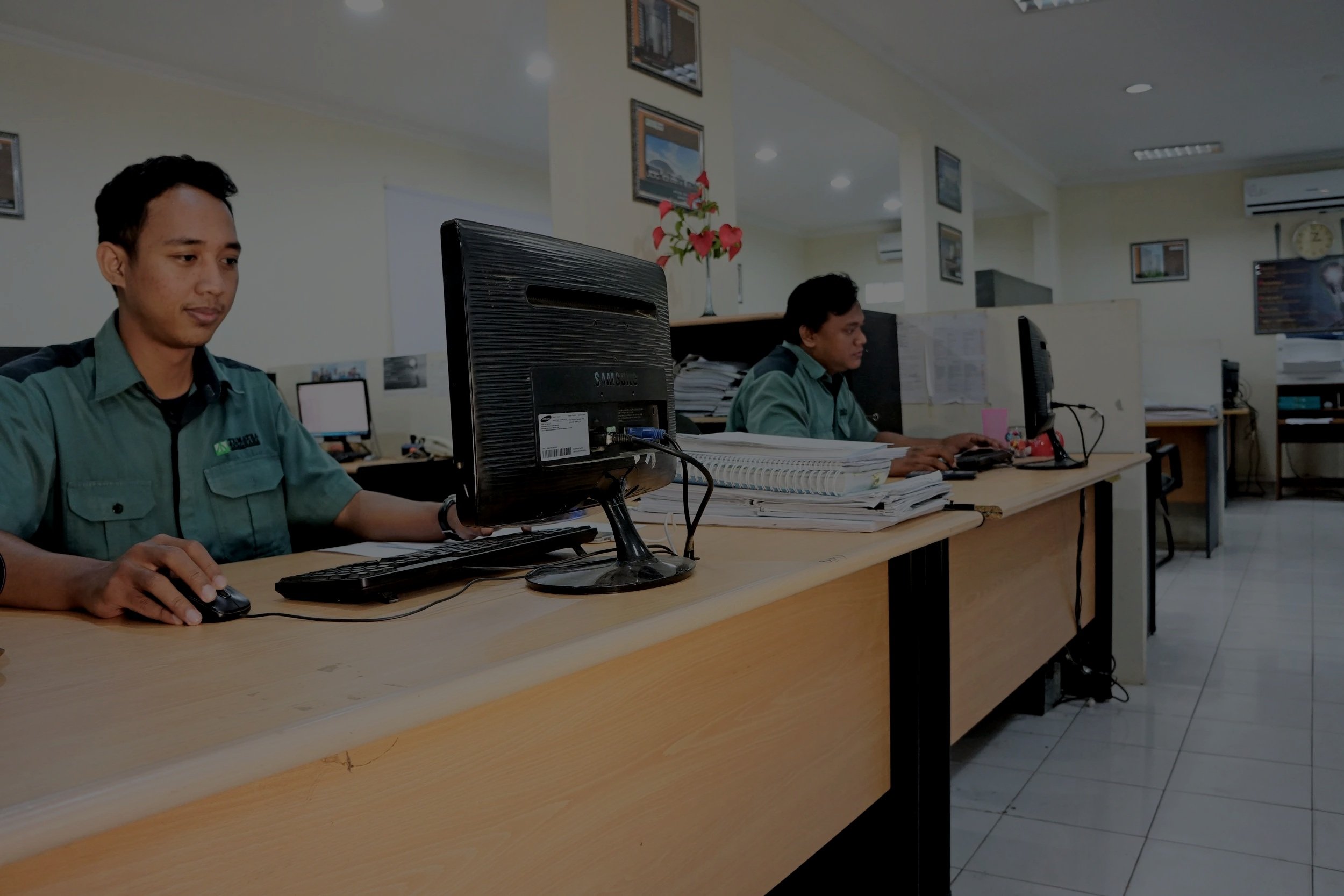 Two men working at desks with computers and stacks of papers in an office with framed pictures on the wall.
