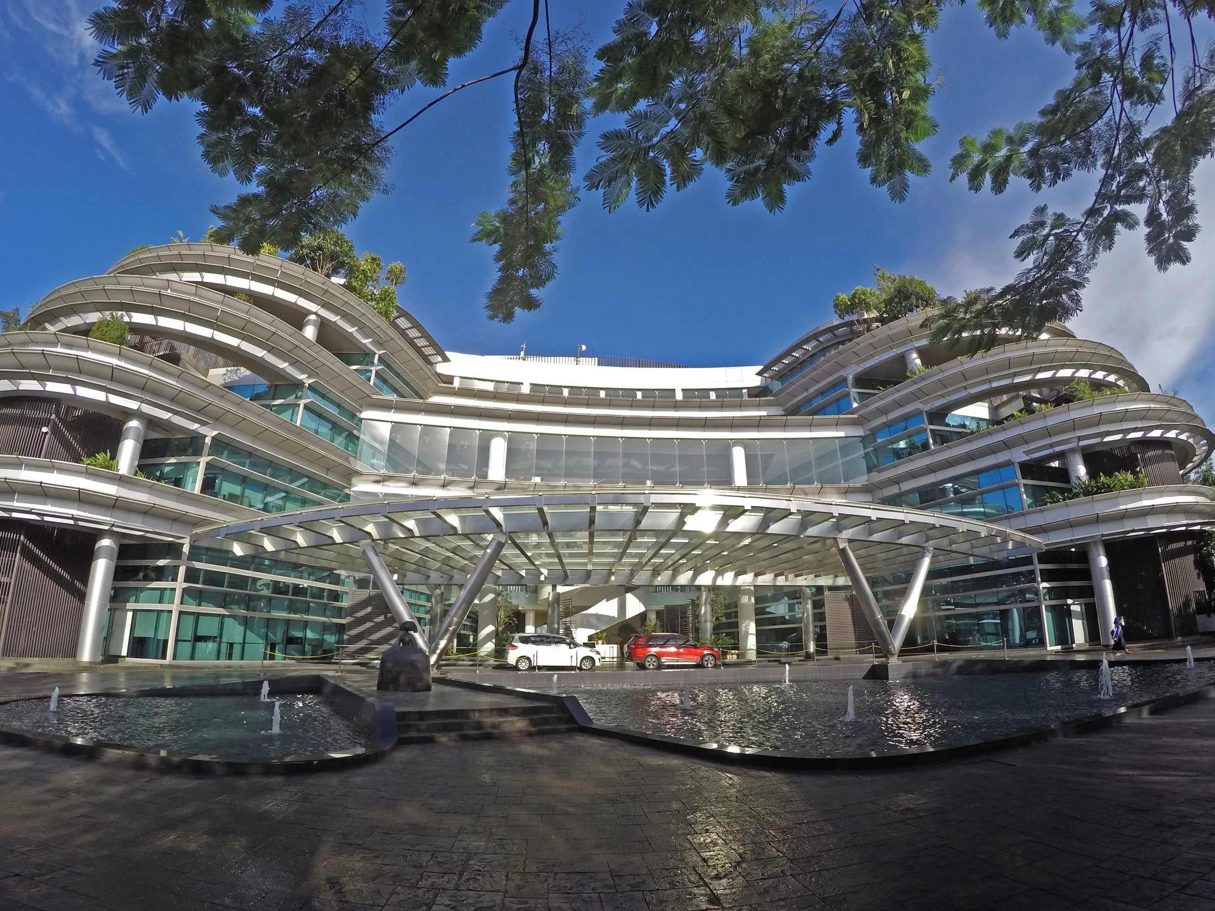 A modern multi-story office building with curved balconies and large glass windows, surrounded by trees and parked cars, with a water fountain in the foreground under a blue sky.