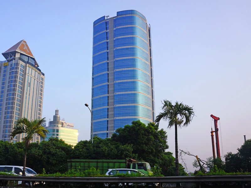 A modern cityscape with tall buildings, including a blue glass skyscraper, and green trees in the foreground.