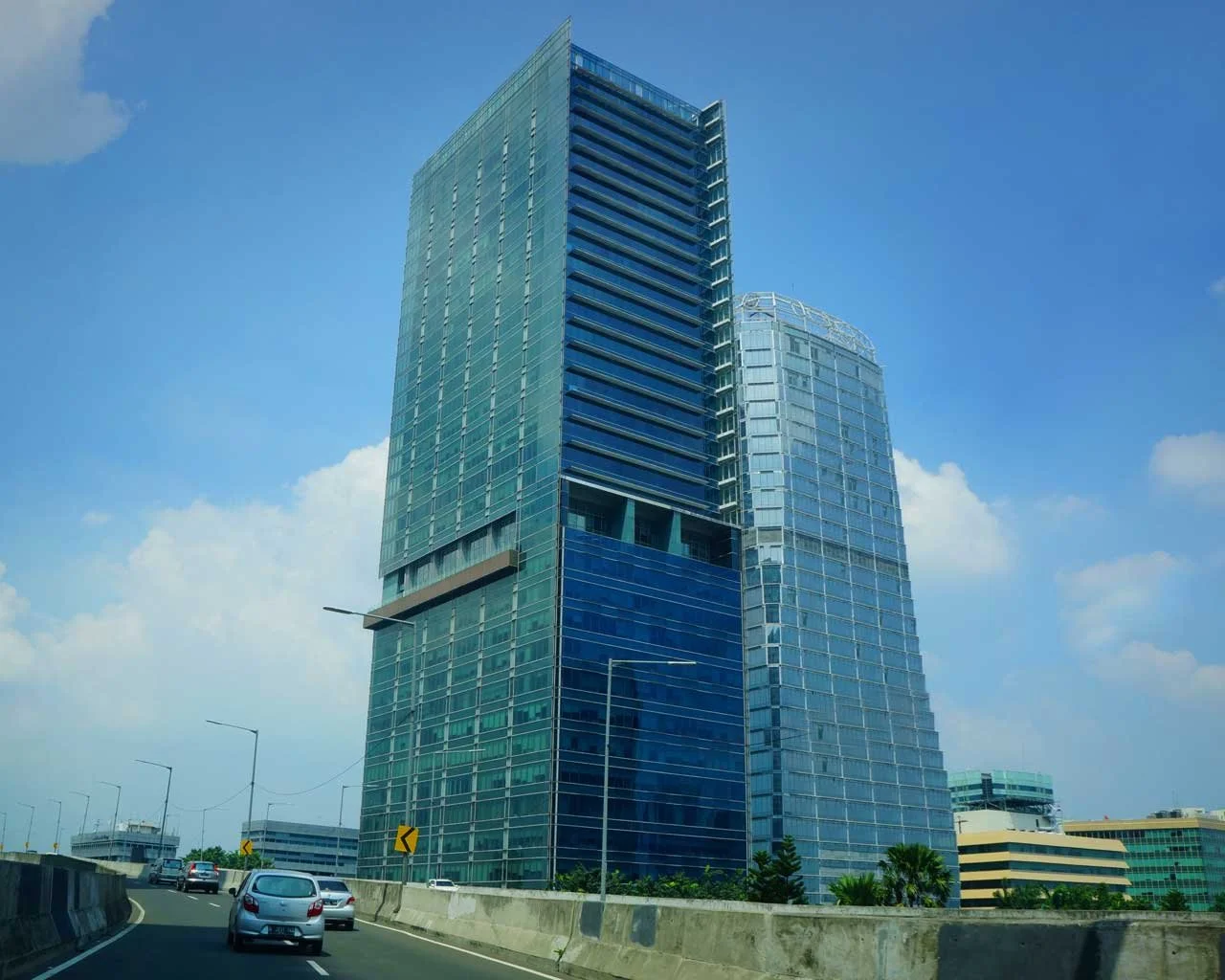 Tall modern glass skyscraper with blue-tinted windows on a highway during daytime, with a few cars and a partly cloudy sky in the background.