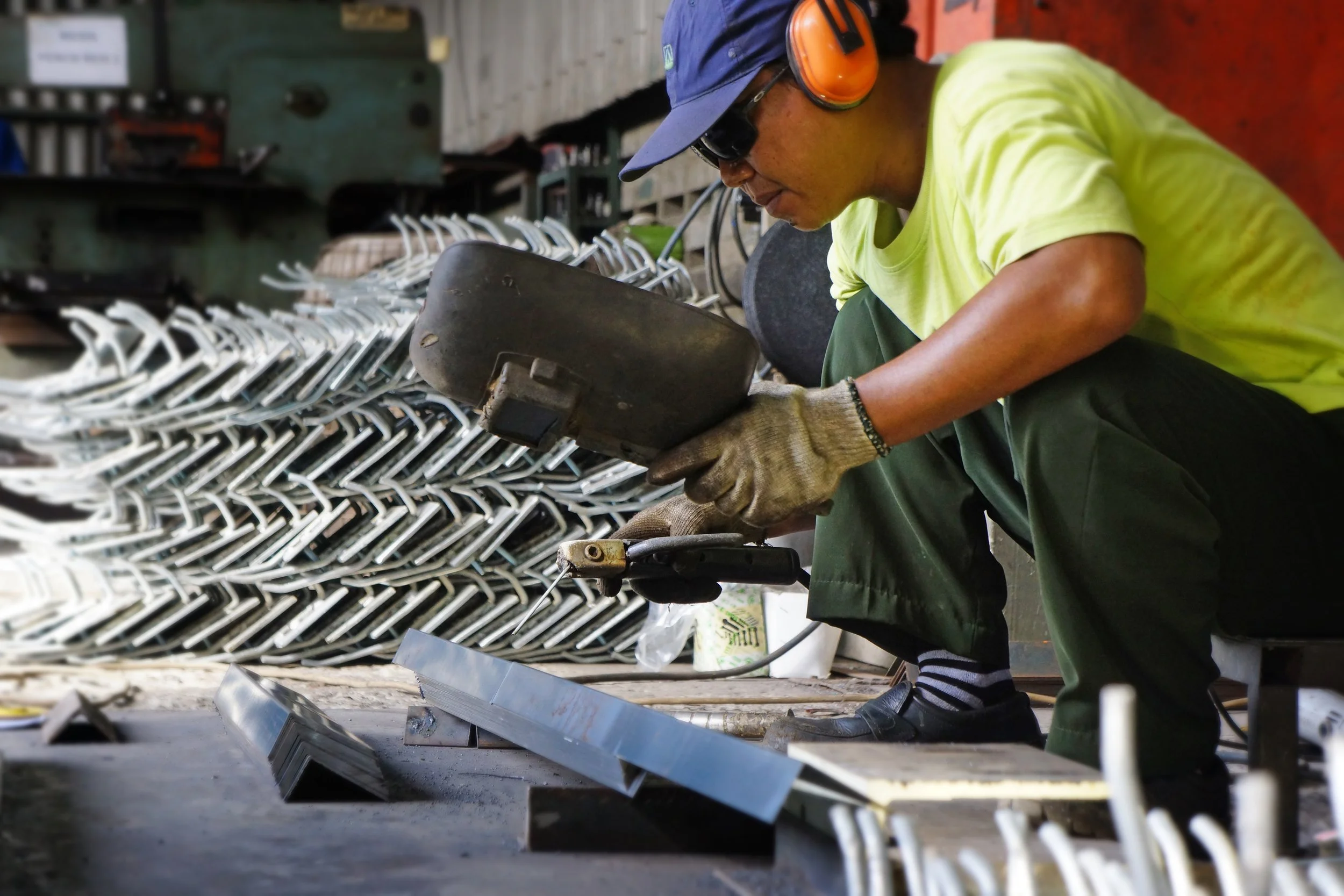 A worker wearing gloves, safety glasses, a blue cap, and ear protection, is welding metal chair frames in a workshop.