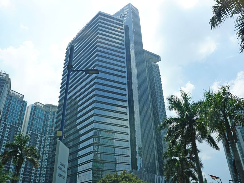 Tall modern glass skyscraper in a city with palm trees in foreground and partly cloudy sky.