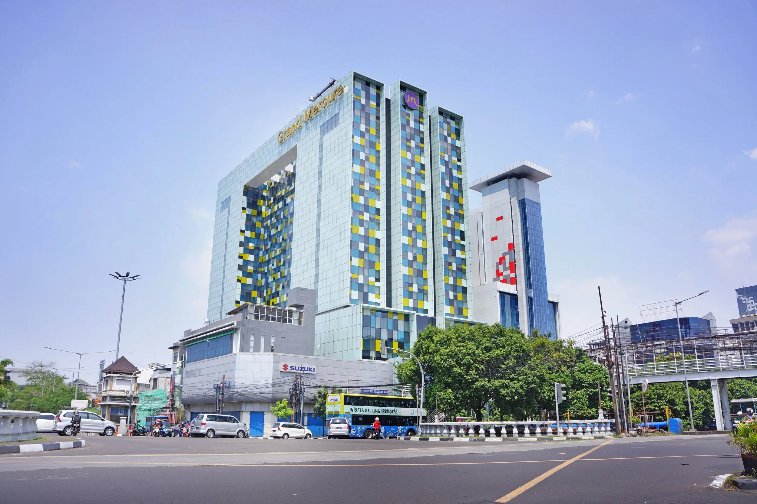 Tall modern building labeled 'Grand Mercure' with colorful glass facade, surrounded by smaller buildings, trees, and a busy street with vehicles and a blue bus.