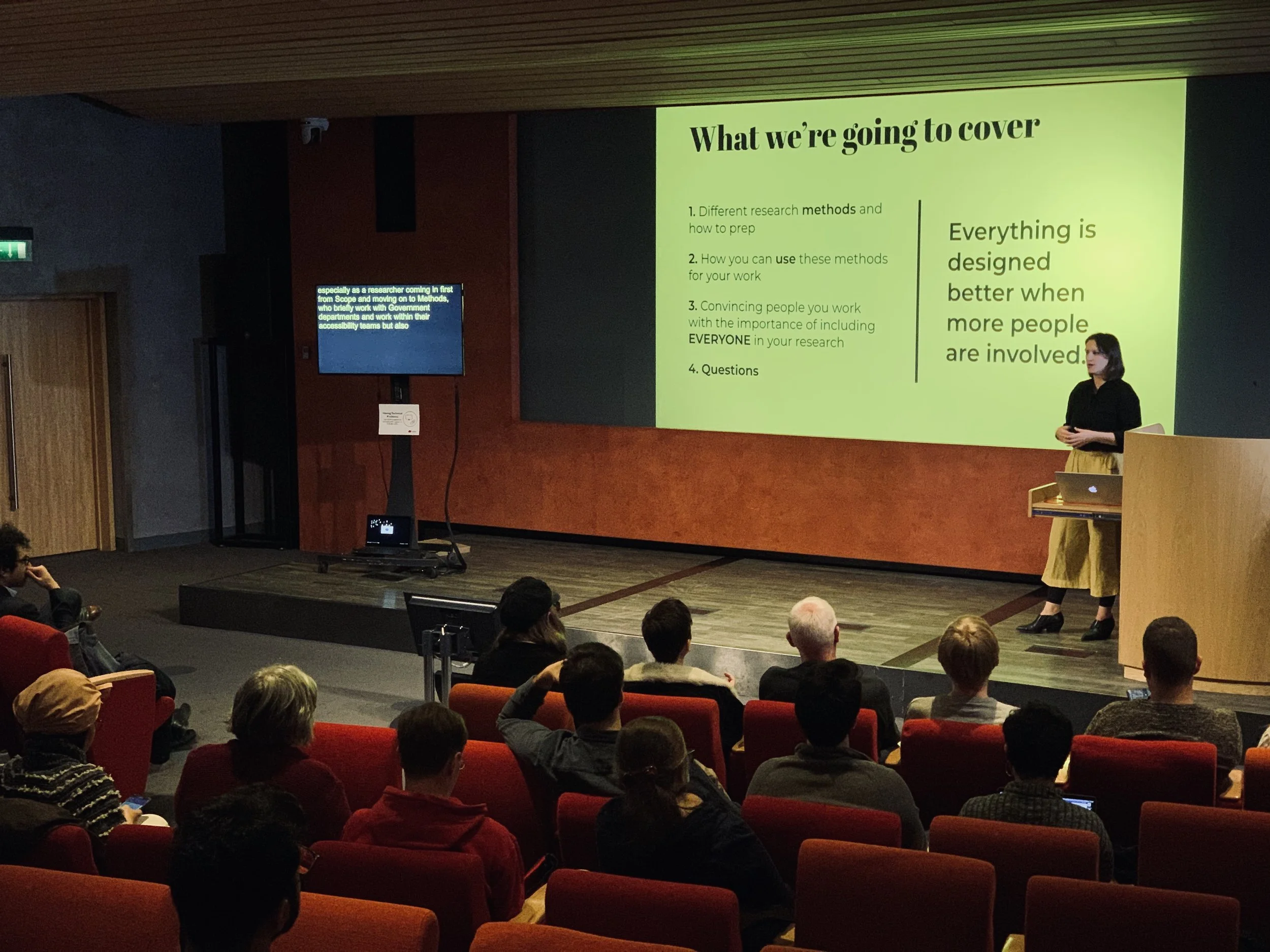 Alex standing next to a screen and in front of an in-person audience giving a presentation