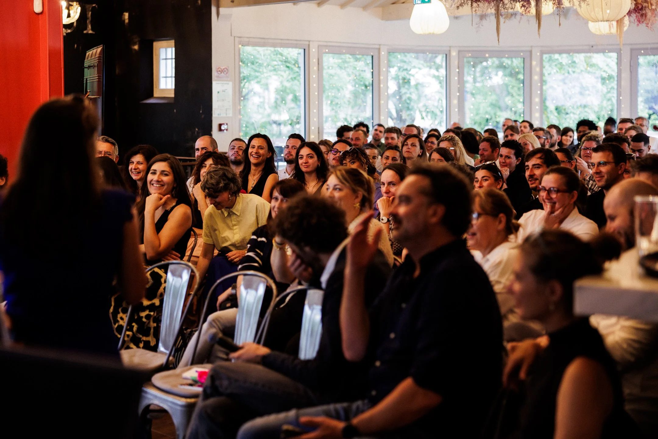 A large group of diverse people sitting in a conference room, listening and smiling during a presentation, with large windows and some hanging lights in the background.