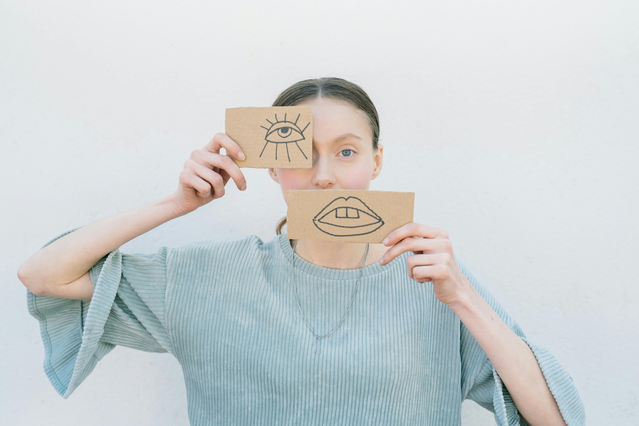Une personne aux yeux bleus tenant deux morceaux de carton avec des dessins représentant un œil et une bouche devant son visage, se tenant devant un fond blanc uni.