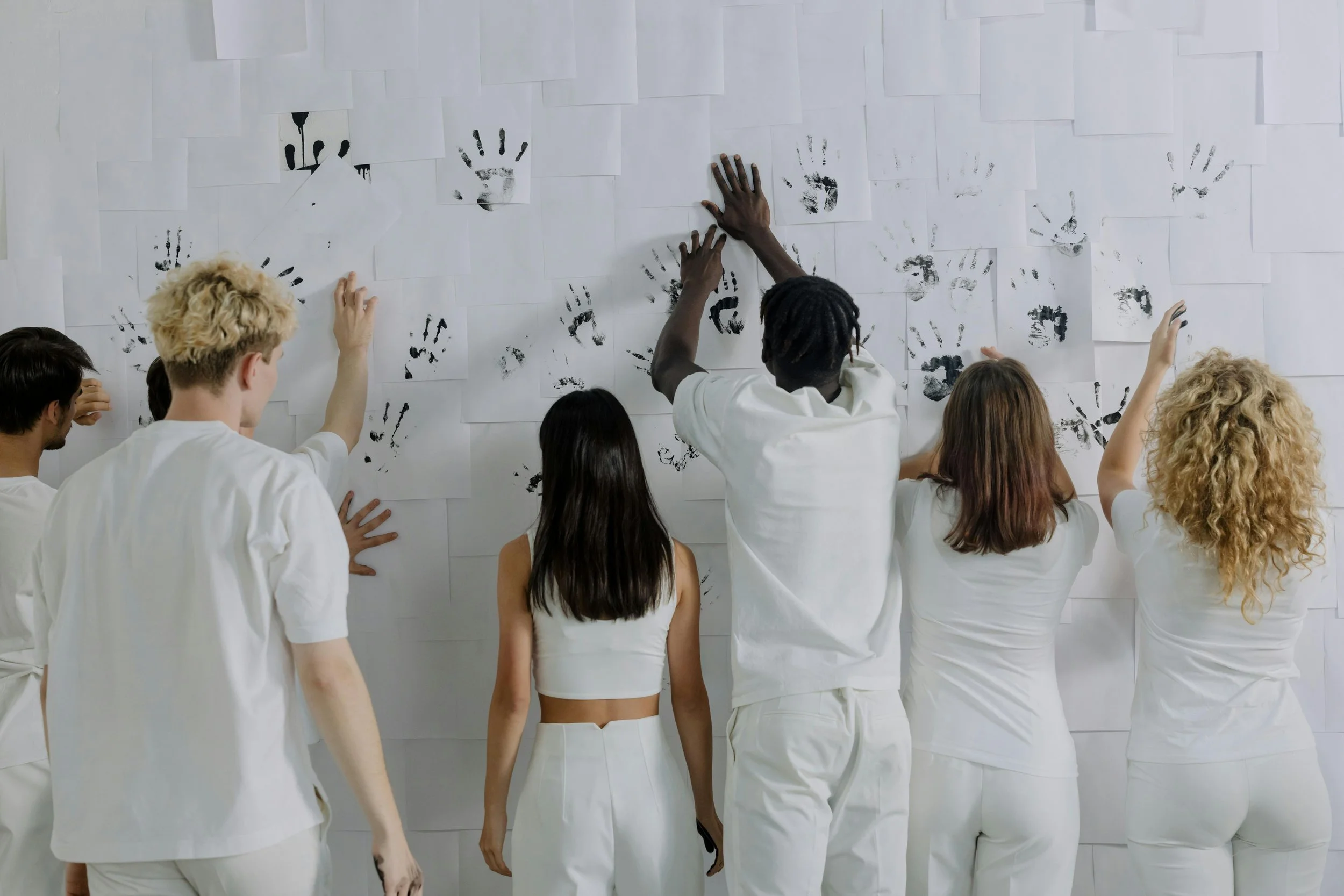 Un groupe de personnes aux profils variés, vêtues de blanc, touchent un mur recouvert d’empreintes de mains.