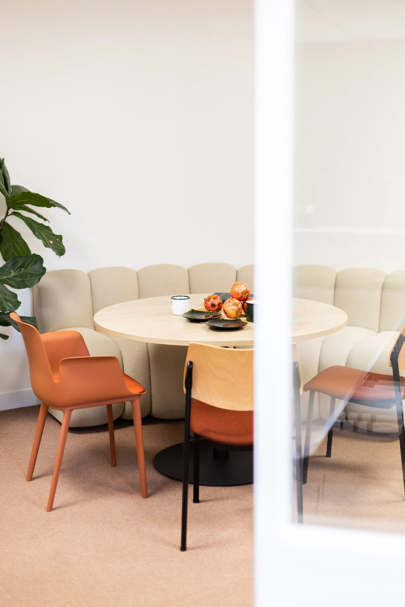 A modern, minimalist dining area with a round light wood table surrounded by chairs in shades of orange, pink, and wood, and a beige curved booth seat in the background. On the table, there is a bowl with pomegranates, a black and a white mug. A large green plant is partially visible on the left side of the image, and the scene is viewed through a partially open white door.
