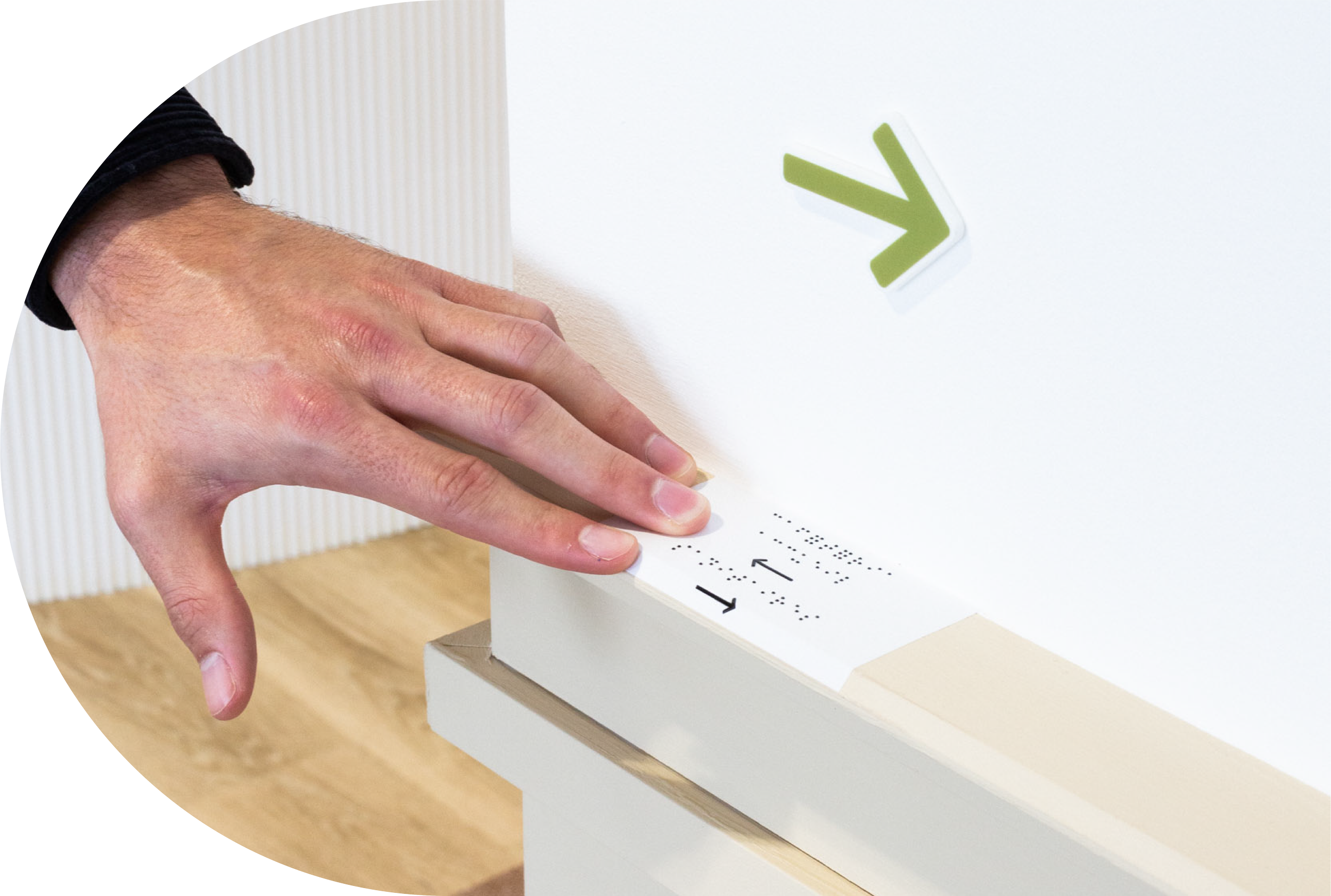 A person with a pinky finger extended touches a white box with black raised dots and an arrow pointing right, on which a green arrow symbol is printed.