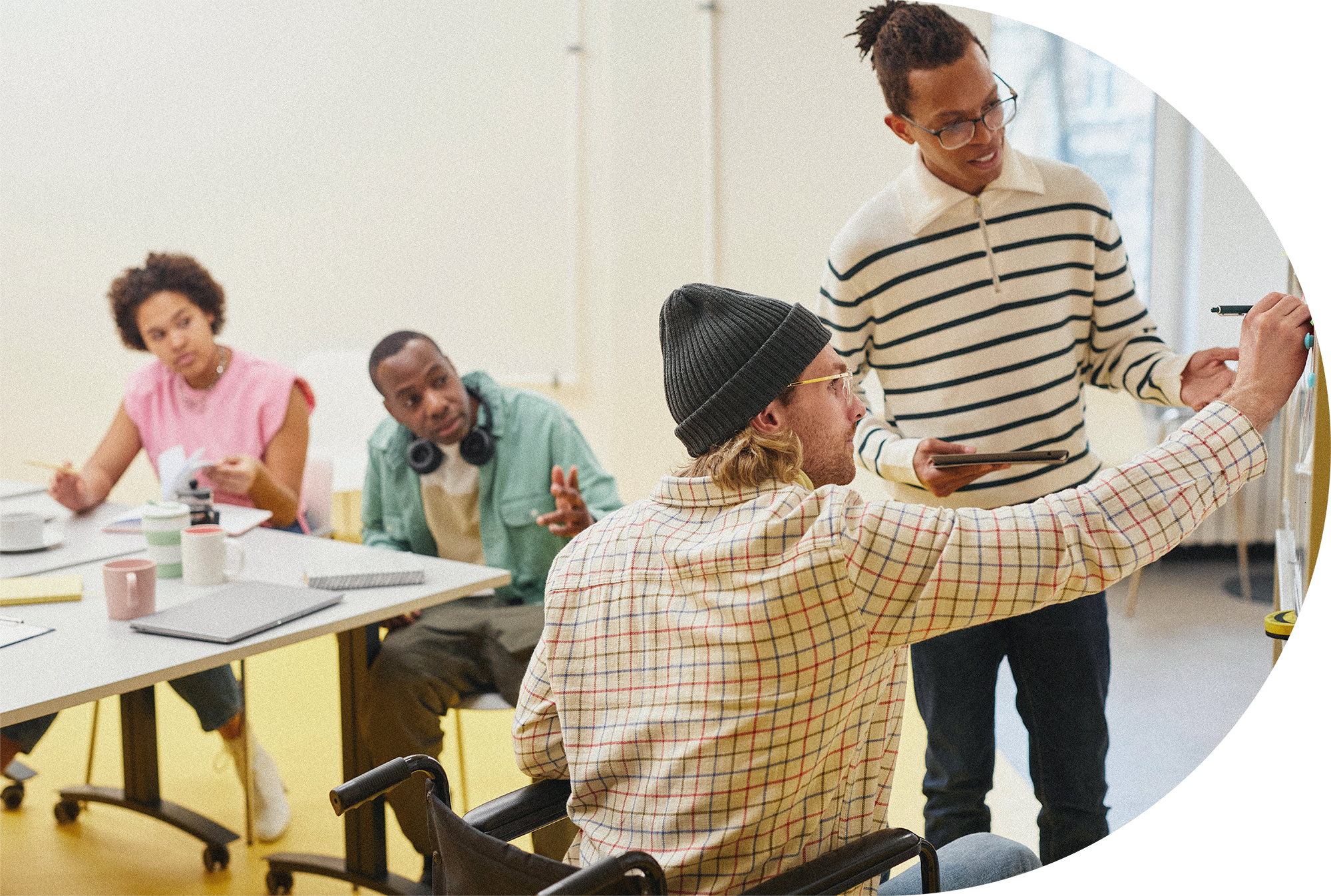 A diverse group of people in a workshop, with one person in a wheel chair writing on a wall, while others are engaging in discussion around a table.