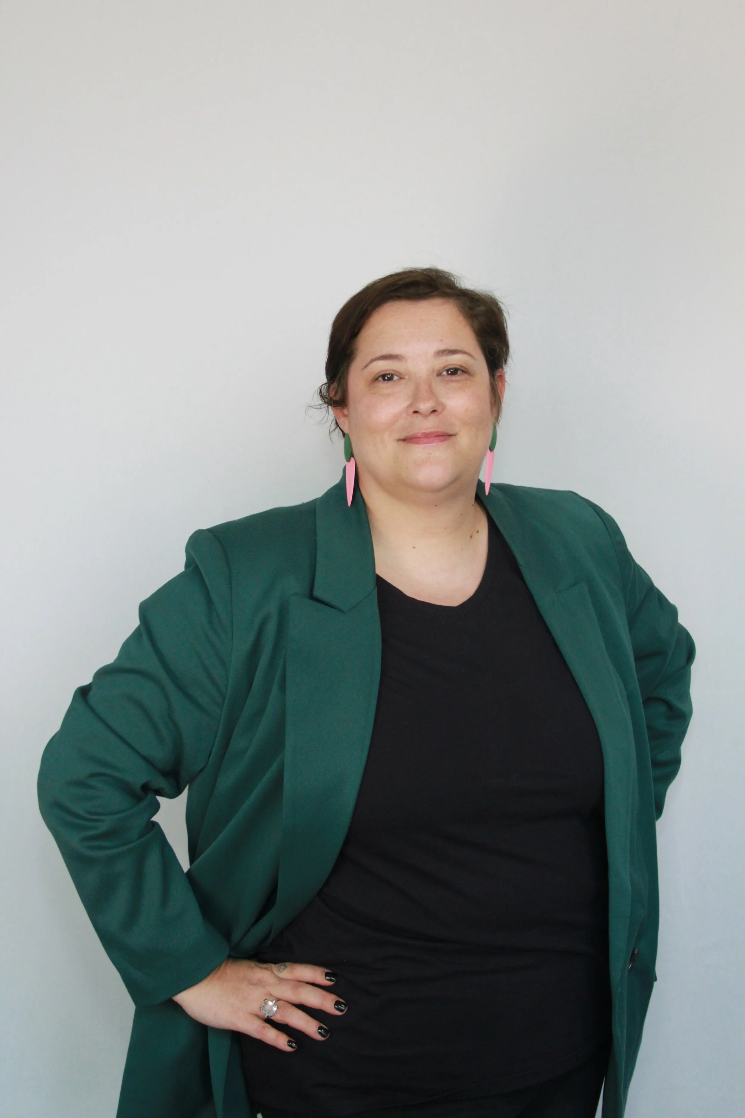Woman with short brown hair wearing green earrings, a black top, and a green blazer, posing with her right hand on her hip, standing against a plain white wall.