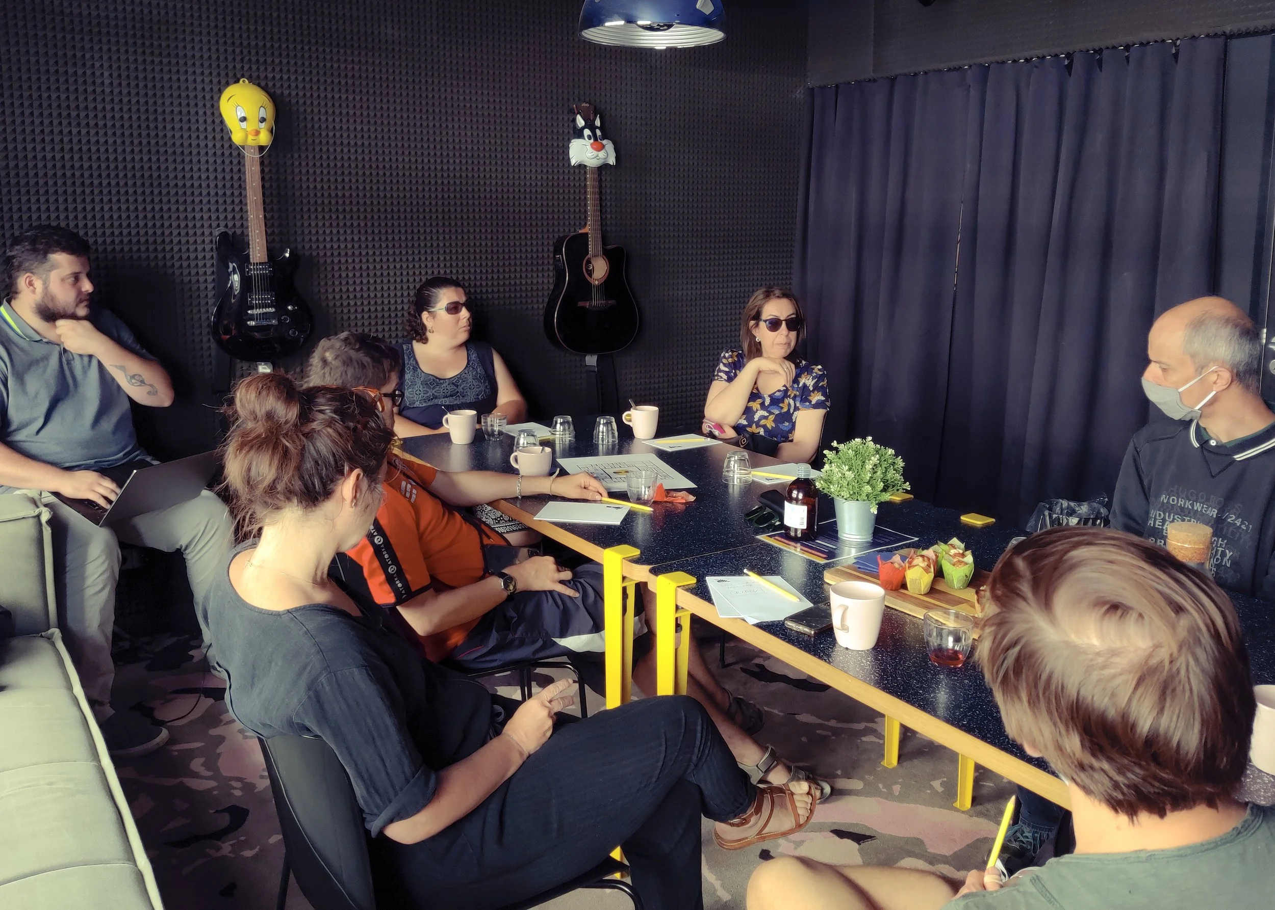 People sitting around a table in a room with black foam soundproofing on the walls, decorated with cartoon character balloons, including a yellow character and a black-and-white character, with drinks and notebooks on the table.