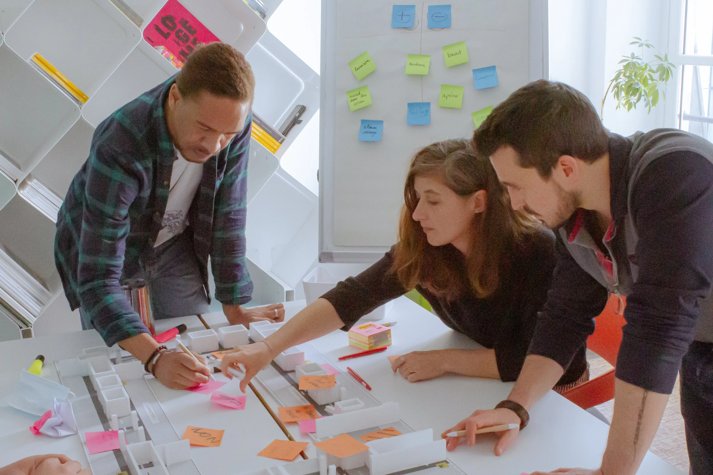 Group of four people gathered around a table working on a project with colorful sticky notes and small white containers, in a bright room with a whiteboard with sticky notes and a window with a plant.