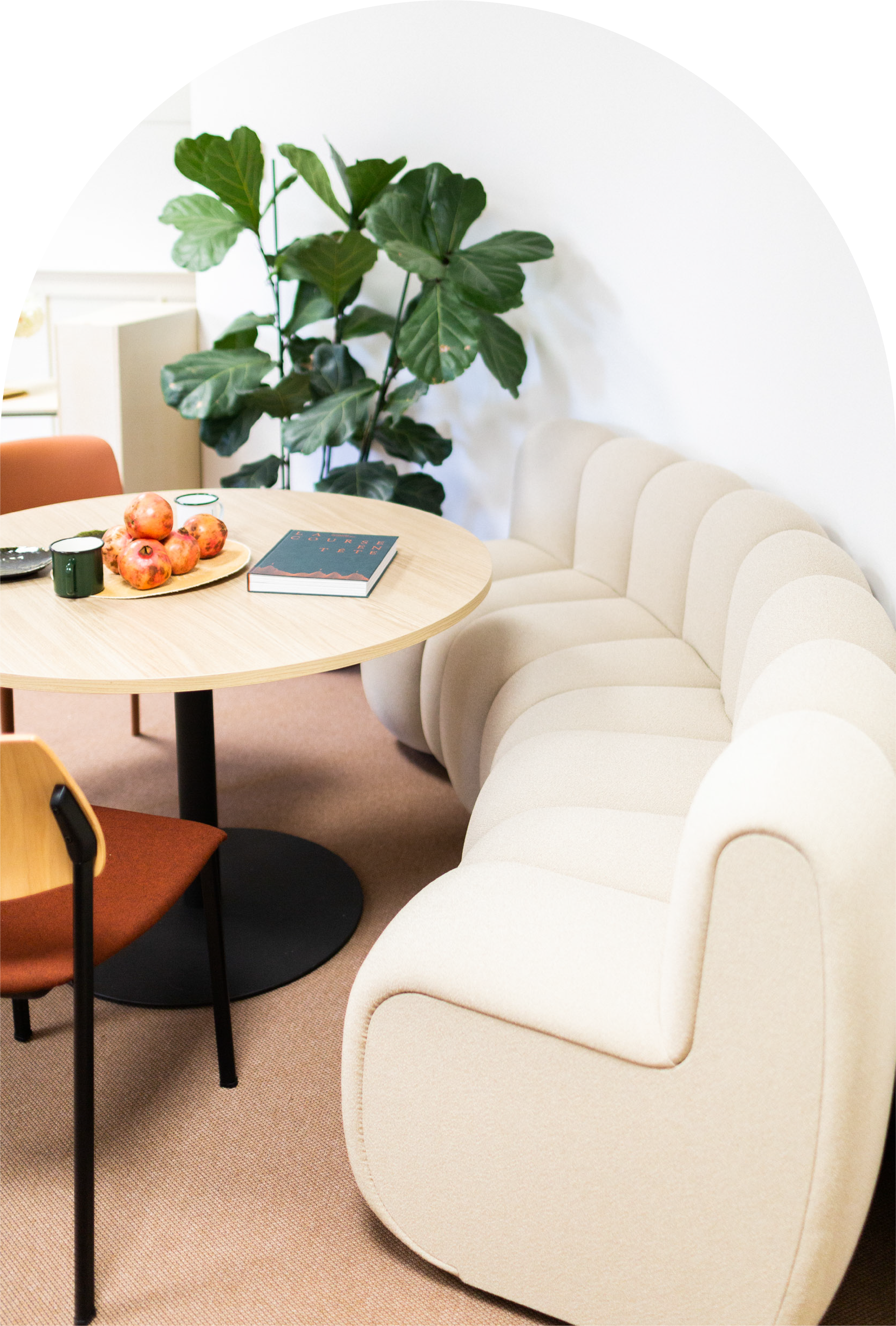 A round wooden table with a bowl of pomegranates, two green cups, and a notebook with a mountain design. Surrounding the table are beige upholstered chairs and an orange chair, with a large green leafy plant in the background against a white wall.