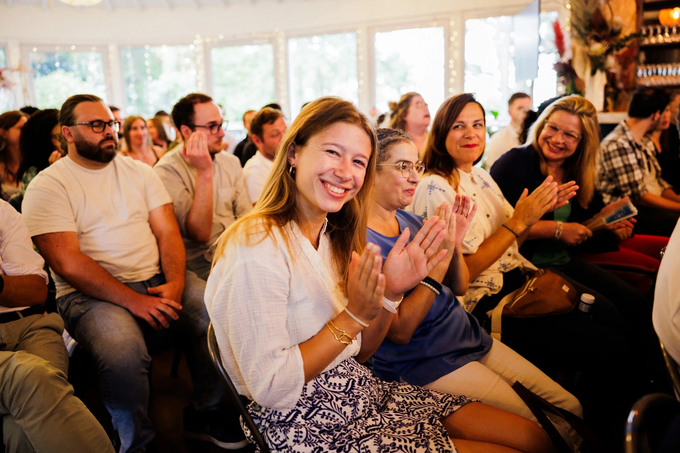 People seated in an audience, clapping and smiling at an event in a bright room with large windows and string lights.