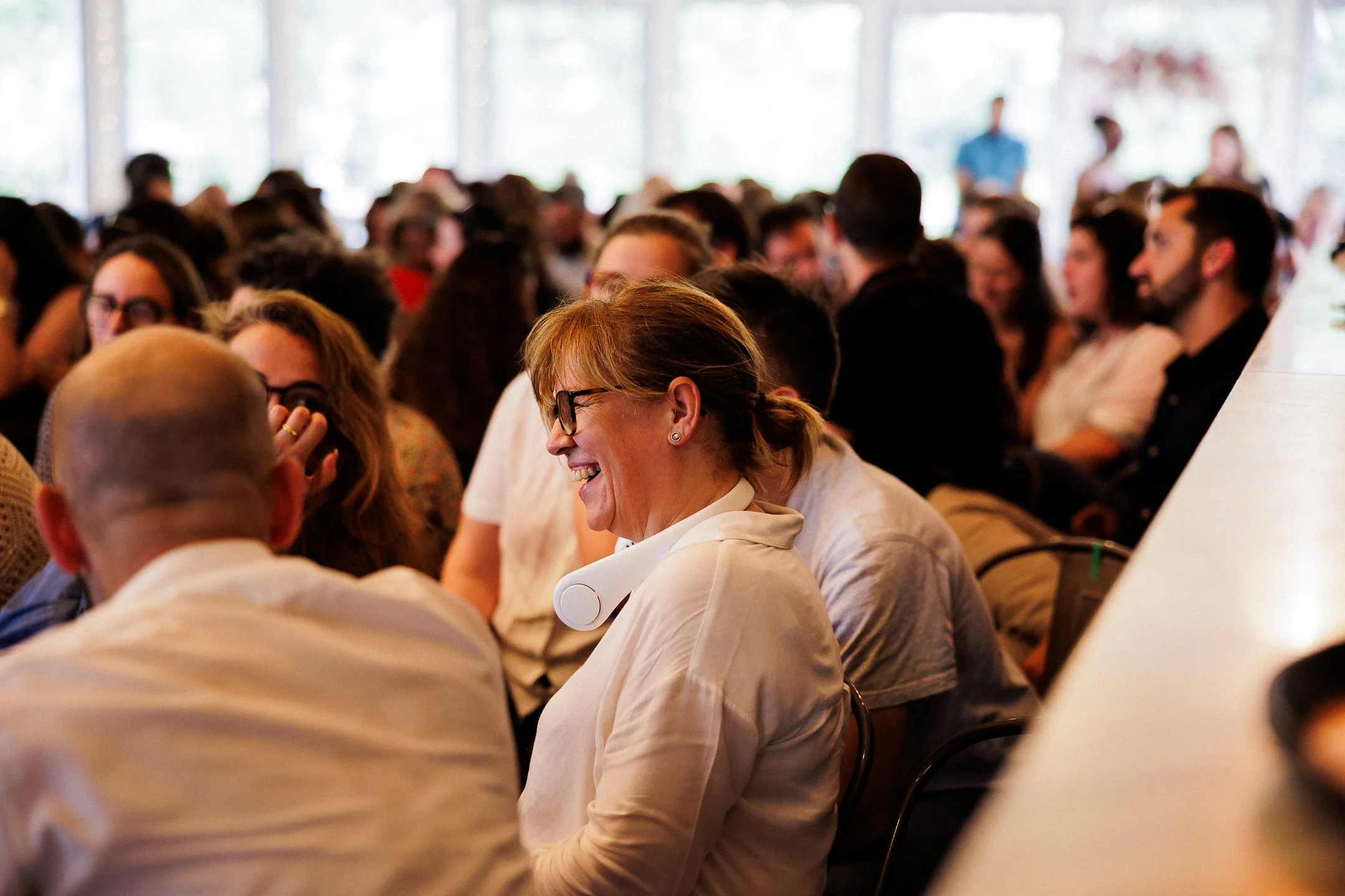 A group of people attending a conference or seminar in a large, bright room with large windows. The focus is on a woman with glasses smiling and engaging with others, while others in the background are listening.