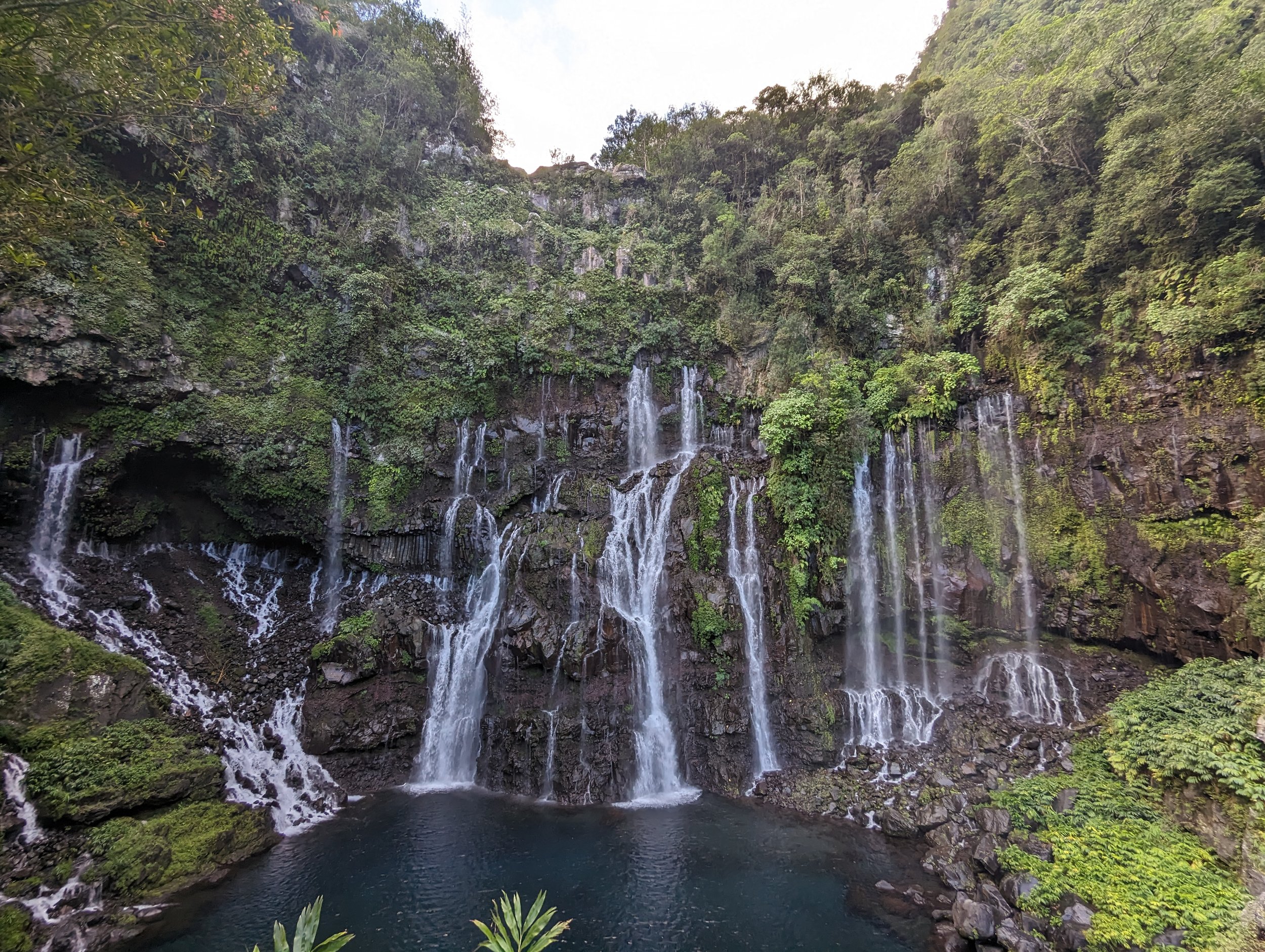Chutes d'eau tombant d'une falaise rocheuse entourée de végétation dense, avec un bassin d'eau en bas.