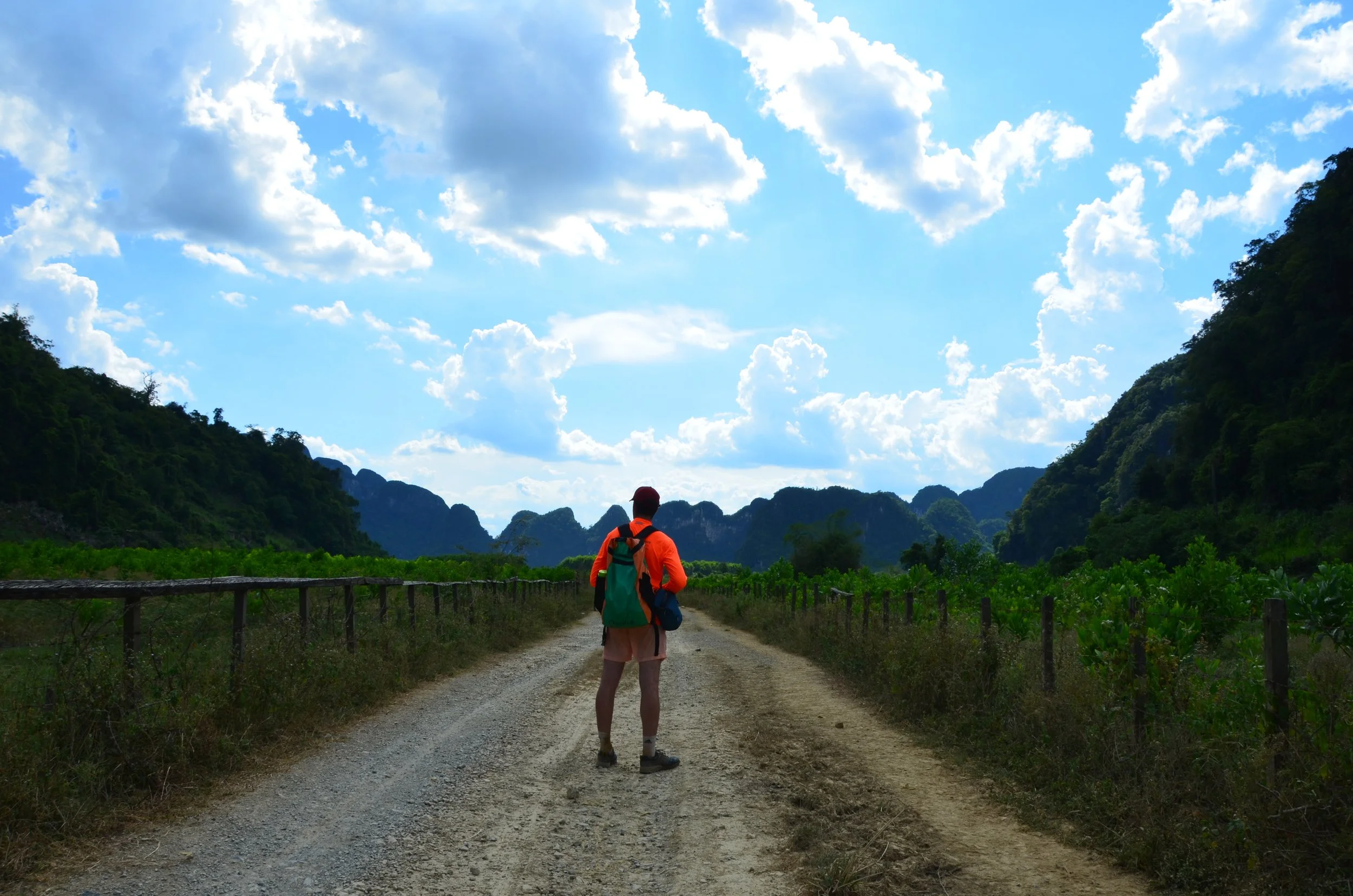 Hiker marchant sur un chemin de terre entouré de végétation, avec montagnes en arrière-plan et un ciel bleu avec des nuages.