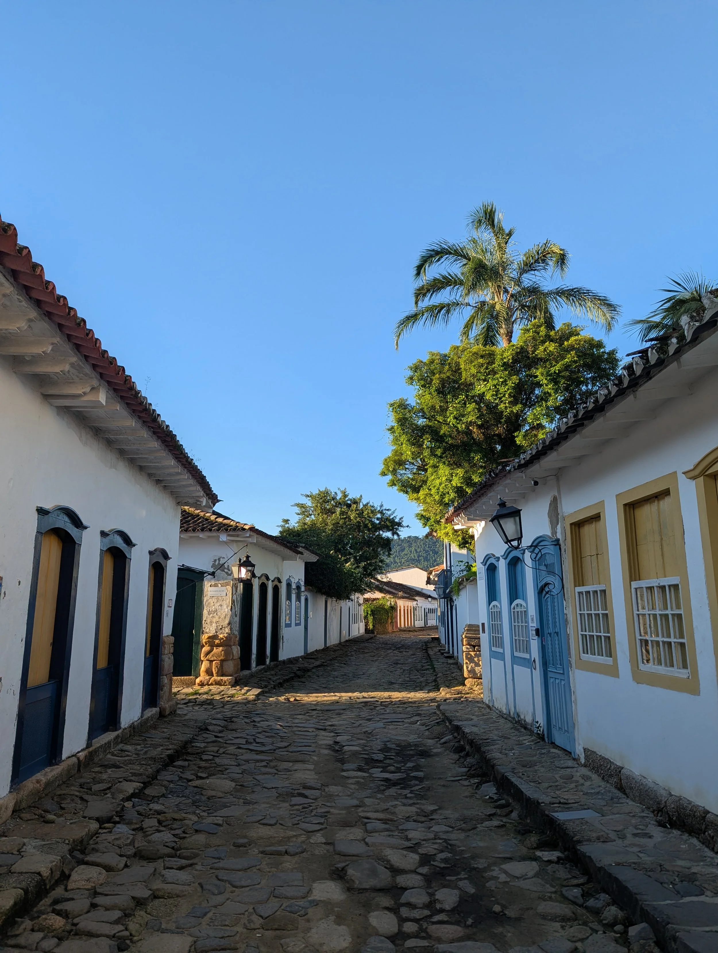vue d'une rue pavée avec des maisons blanches, des fenêtres avec volets, et des arbres en arrière-plan, sous un ciel bleu