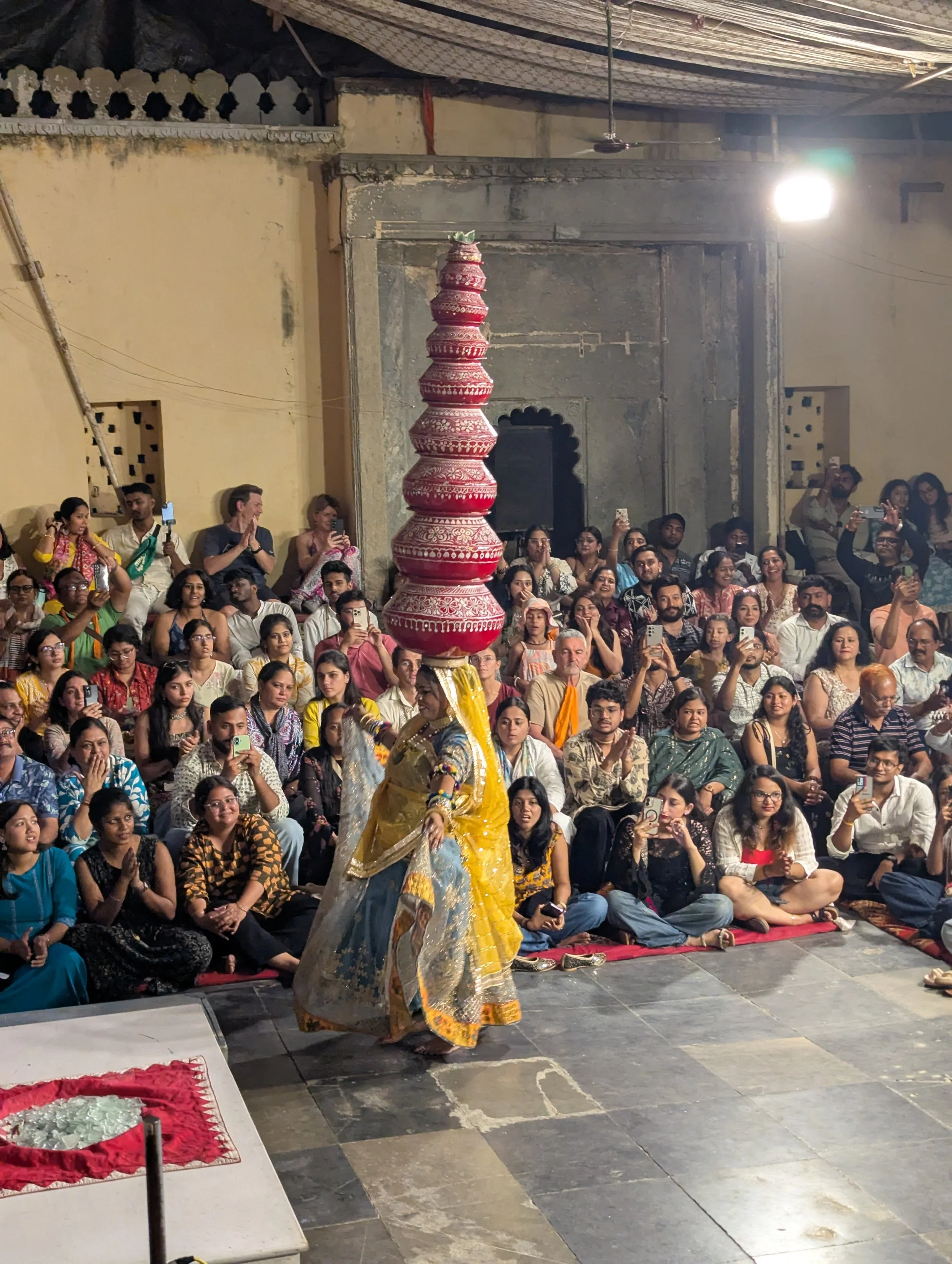 Une femme en costume traditionnel indien avec un grand toupé enroulé, danse devant une foule assise et debout dans un lieu culturel. La foule regarde et prend des photos de la performer.