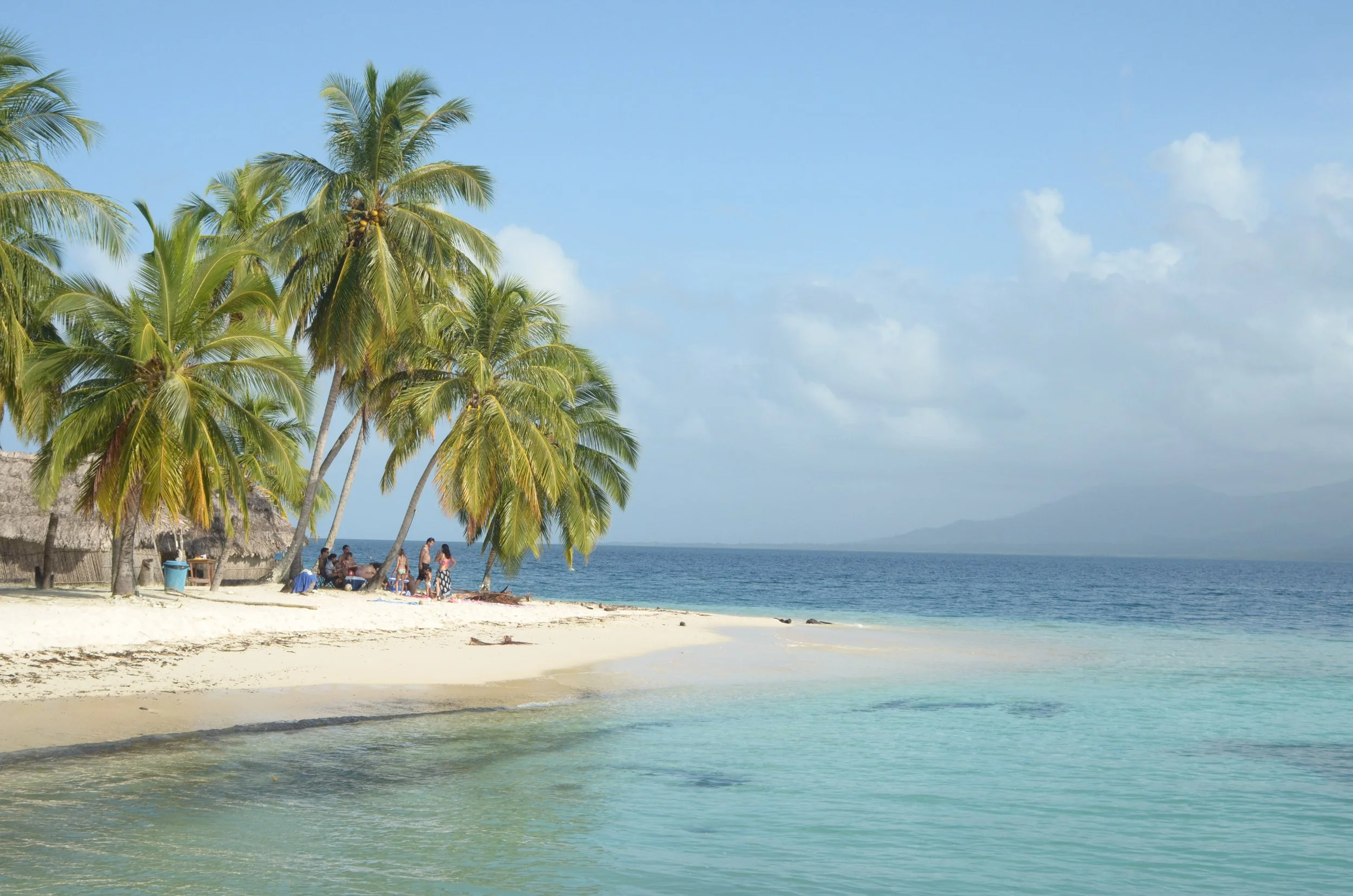 Plage de sable avec des palmiers, une petite cabane et des personnes assises à proximité, vue sur la mer et les montagnes au loin, ciel partiellement nuageux.