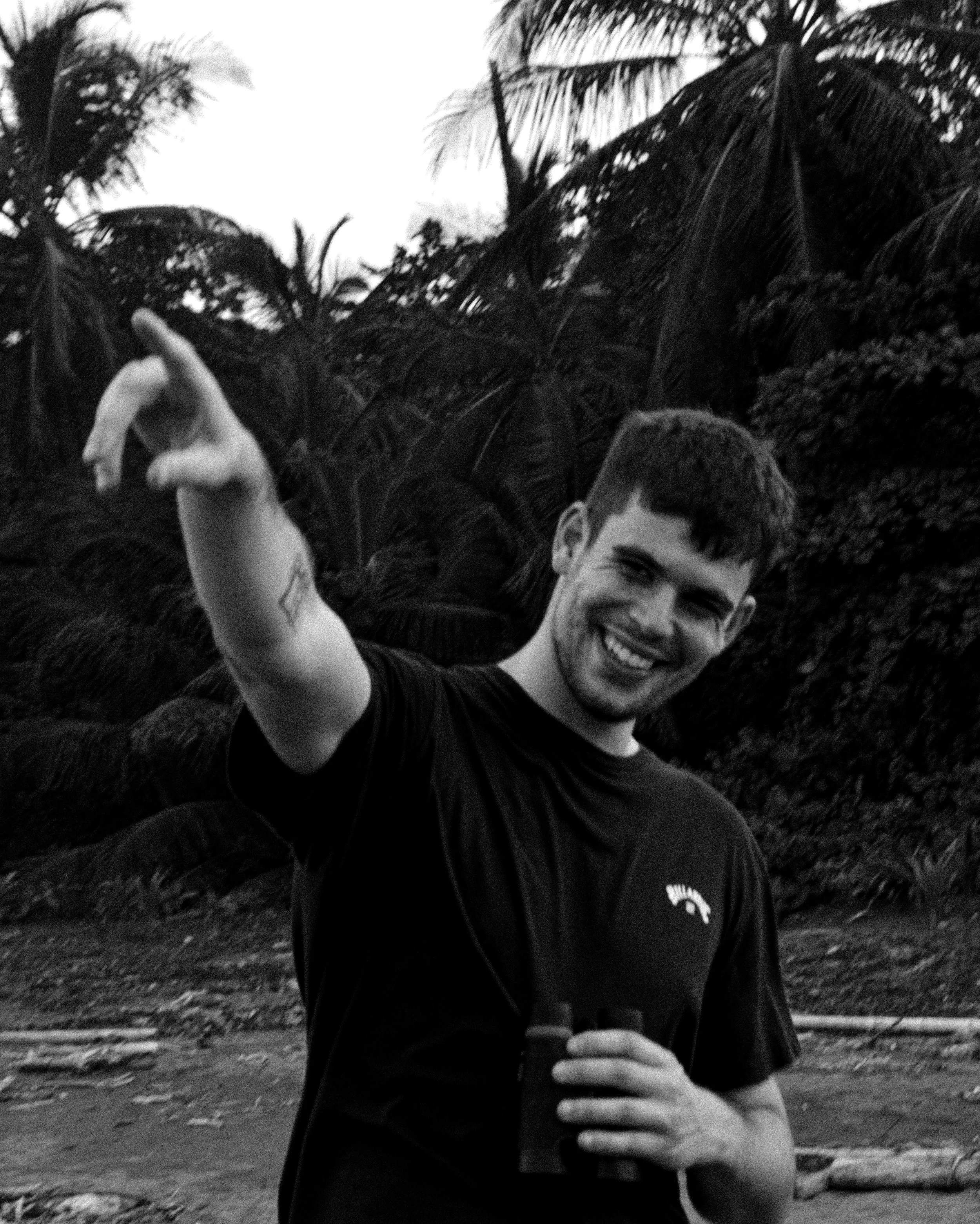 Jeune homme, portant un t-shirt noir, pointant vers l'appareil photo avec une main, en arrière-plan des palmiers et une nature tropicale.