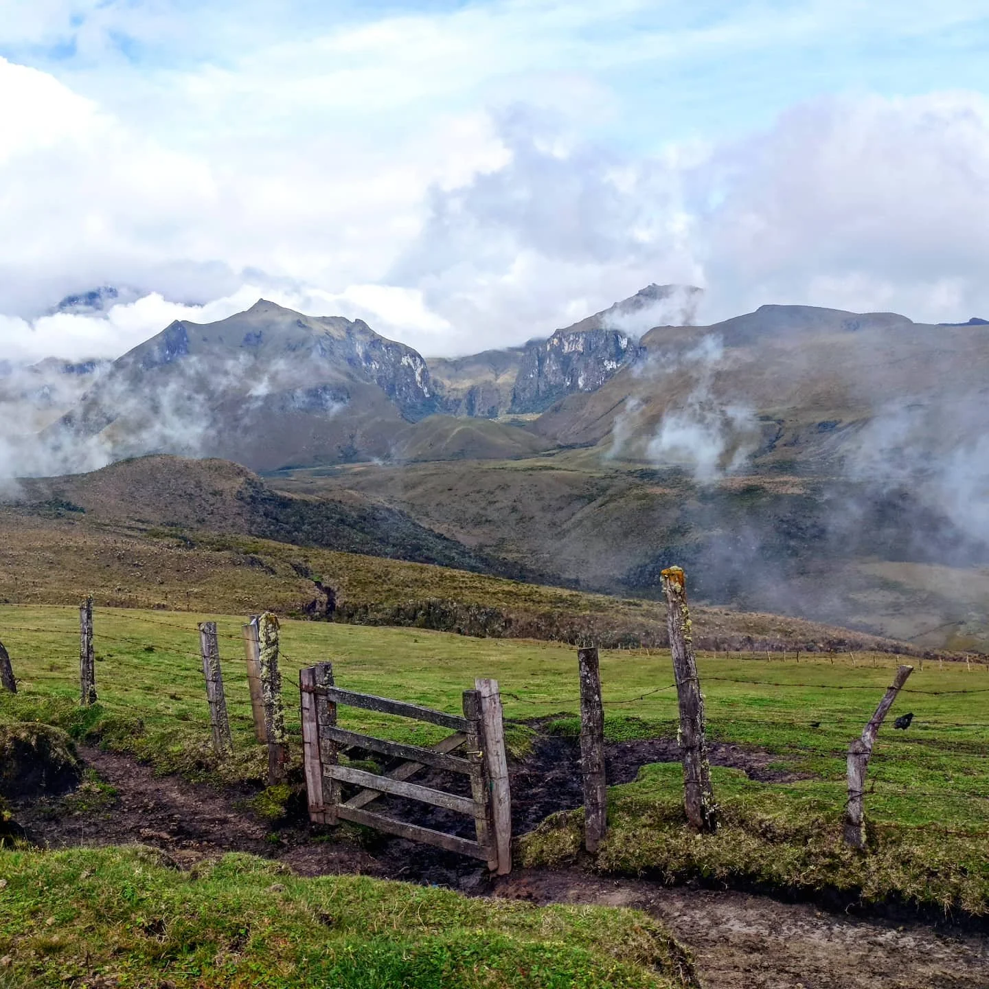 Paysage de montagnes avec brume et vapeur, clôture en bois endommagée au premier plan, terrain verdoyant.