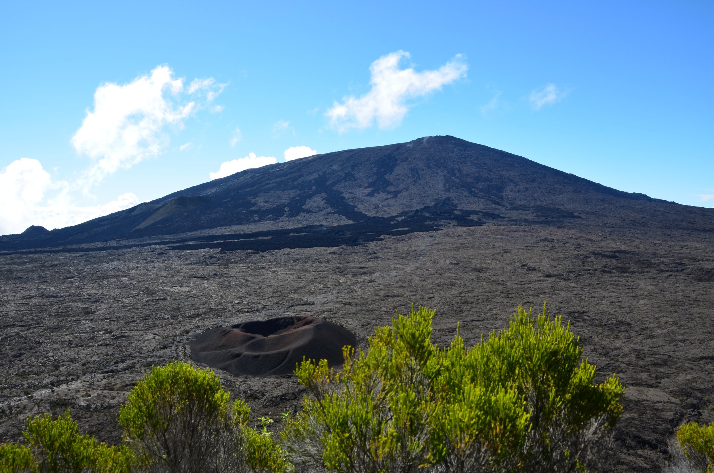 Vue d'un volcan actif avec un cratère au premier plan, entouré de végétation verte, sous un ciel bleu avec quelques nuages.