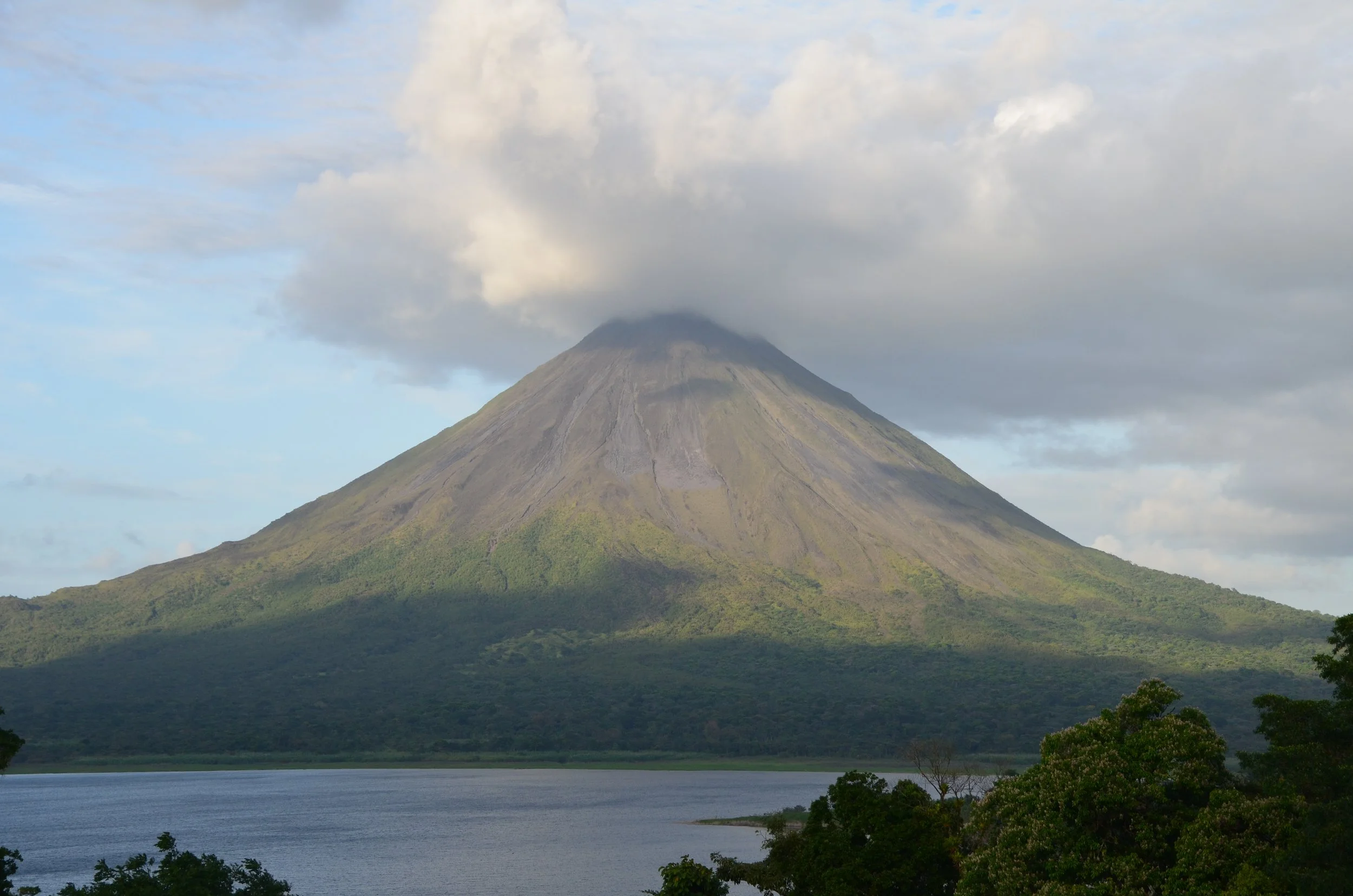 Un volcan avec un sommet en forme de cône, partiellement couvert de nuages, et entouré de verdure et d'un lac au premier plan.