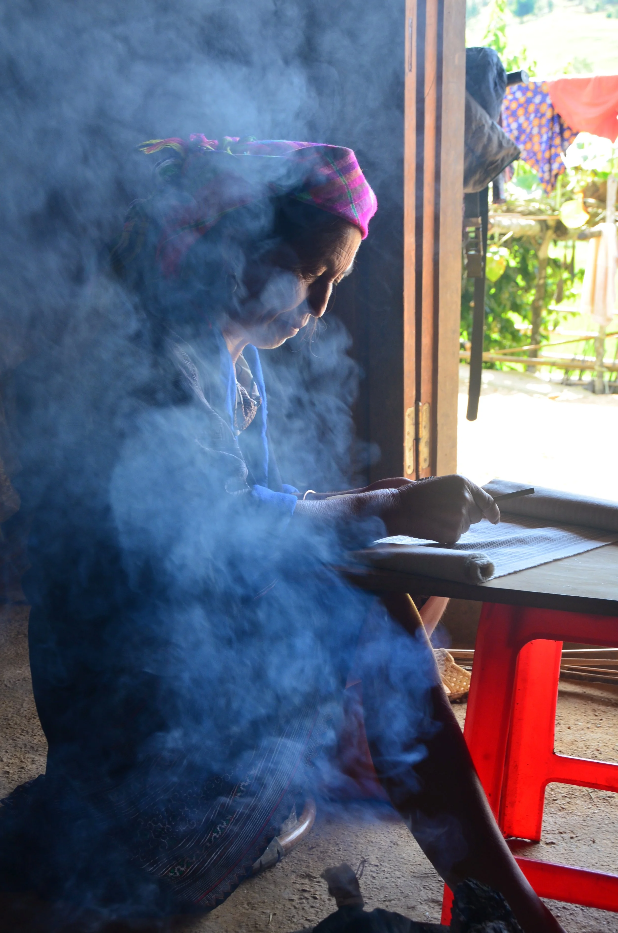Une femme assise à une table, écrivant ou lisant avec de la fumée ou de la brume qui l'entoure, dans un environnement intérieur avec une ouverture vers l'extérieur ensoleillé.