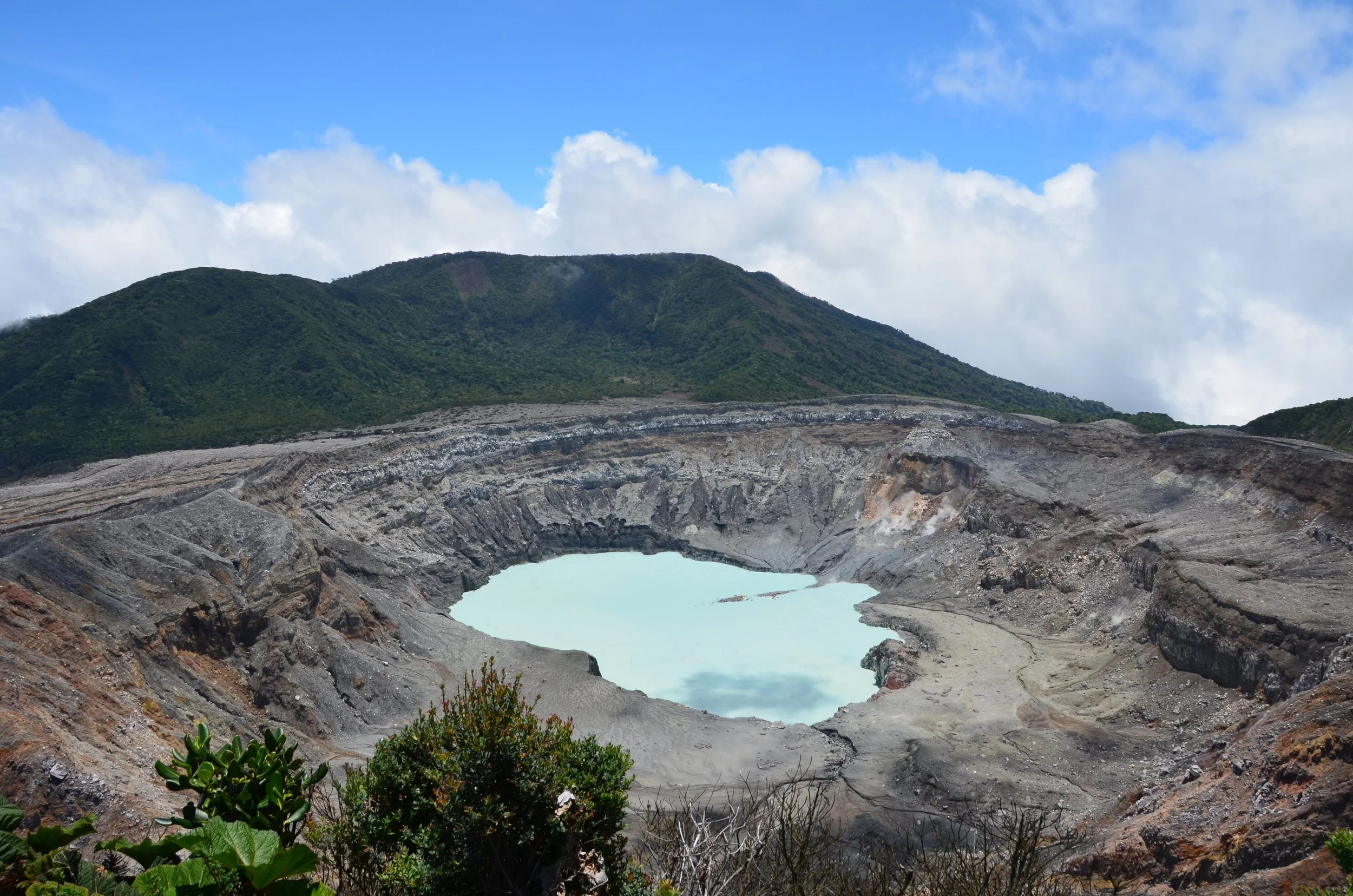 Vulcano avec un cratère rempli d'eau turquoise, entouré de pentes rocheuses et montagne verte en arrière-plan