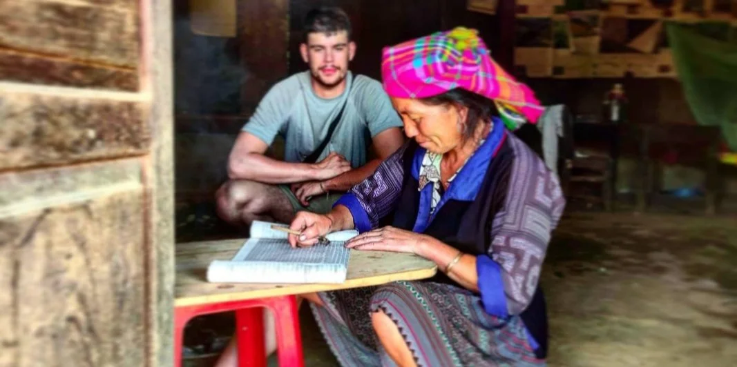 Une femme âgée portant un turban coloré écrit dans un livre posé sur une table tandis qu'un jeune homme la regarde, dans un intérieur en bois.