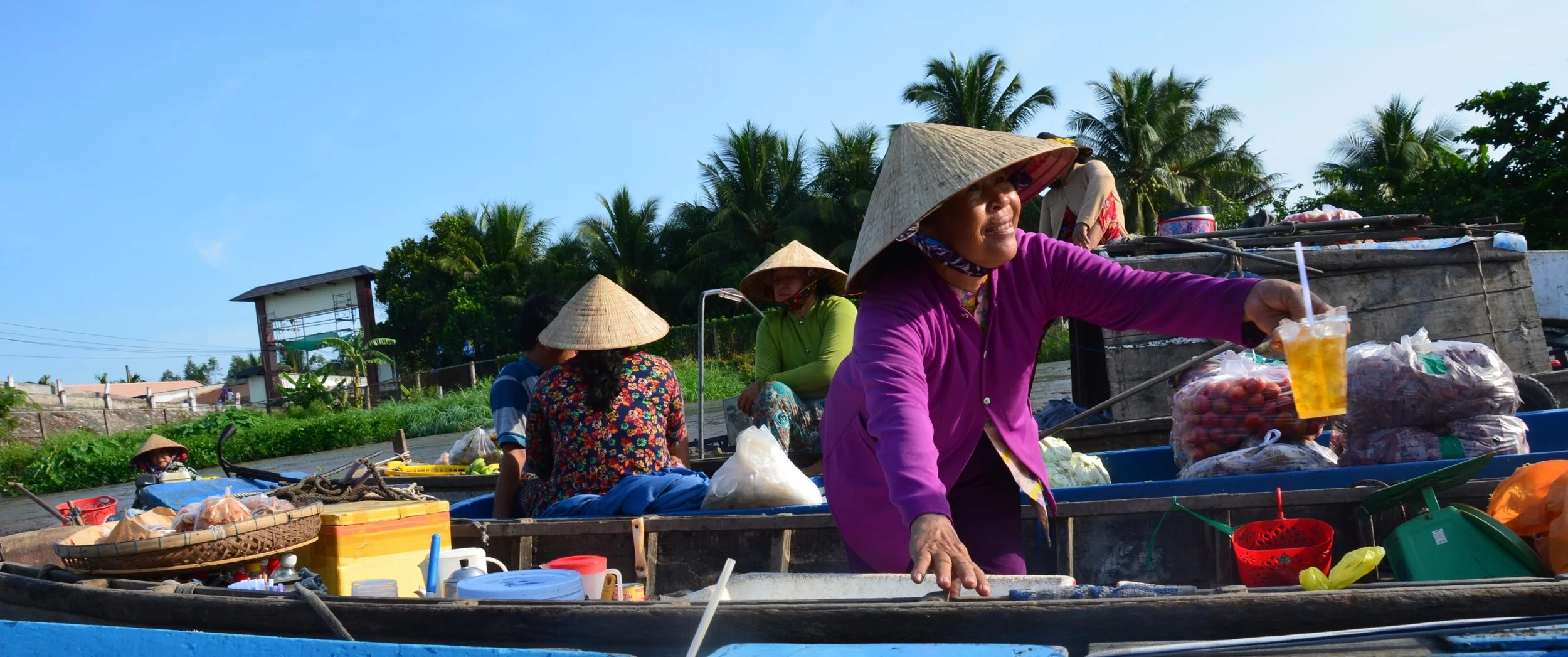 Marché flottant avec femmes portant des chapeaux coniques en vendant des marchandises et boissons à bord de bateaux en bois, avec un ciel bleu et des palmiers en arrière-plan.