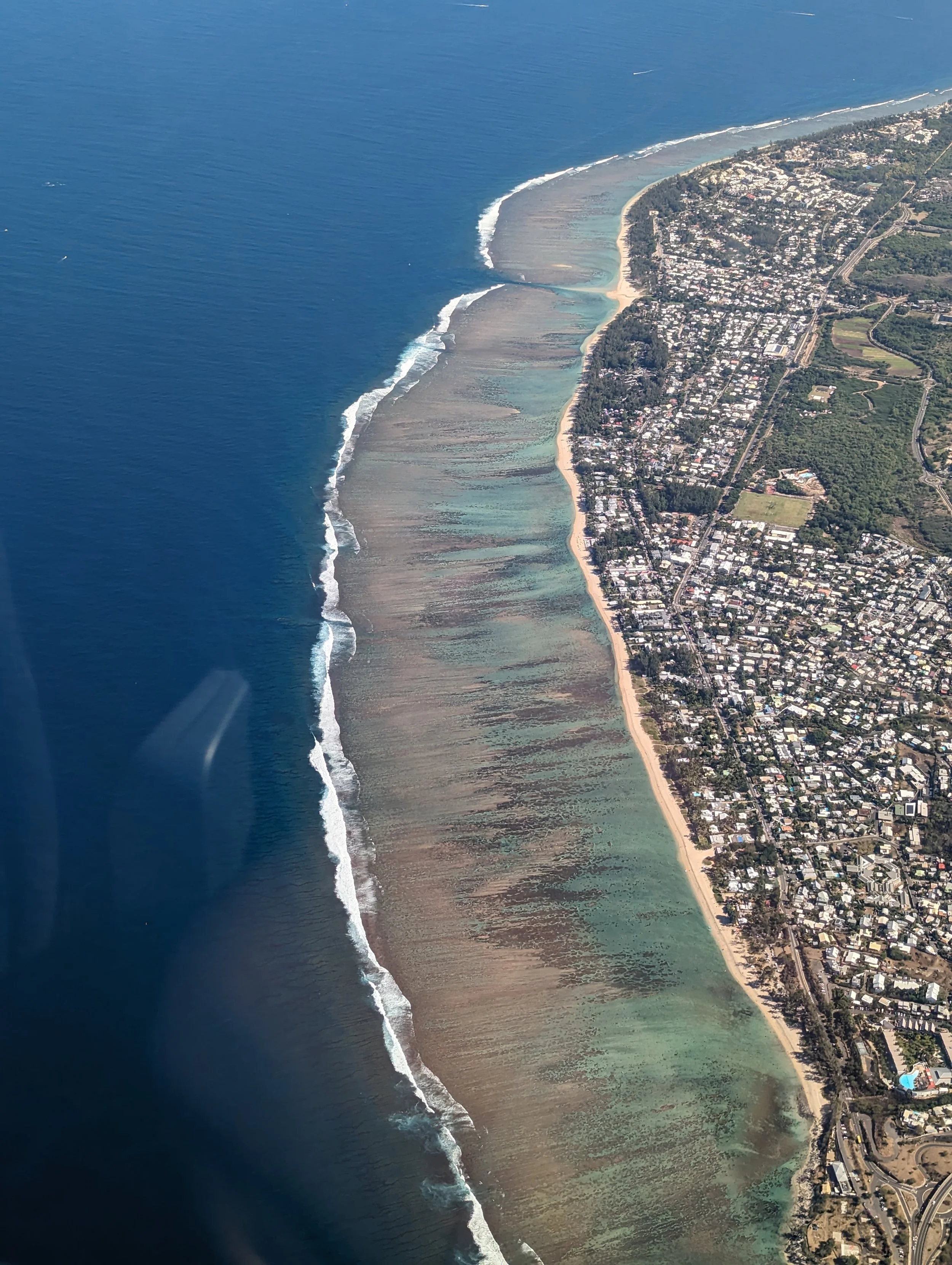 Vue aérienne d'une côte avec plage, eaux turquoise, vagues et zone de marée, bordée par une ville avec bâtiments résidentiels et zones vertes.
