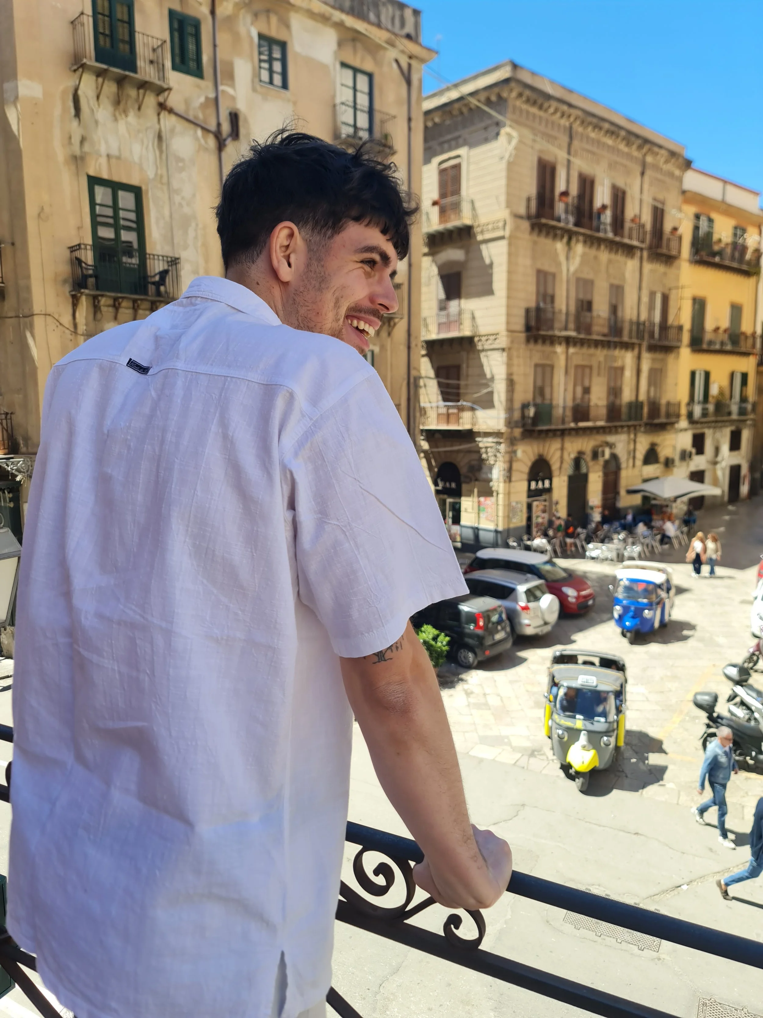 Jeune homme souriant, portant une chemise blanche, regarde par-dessus une balustrade, avec des bâtiments anciens et une rue animée en arrière-plan.