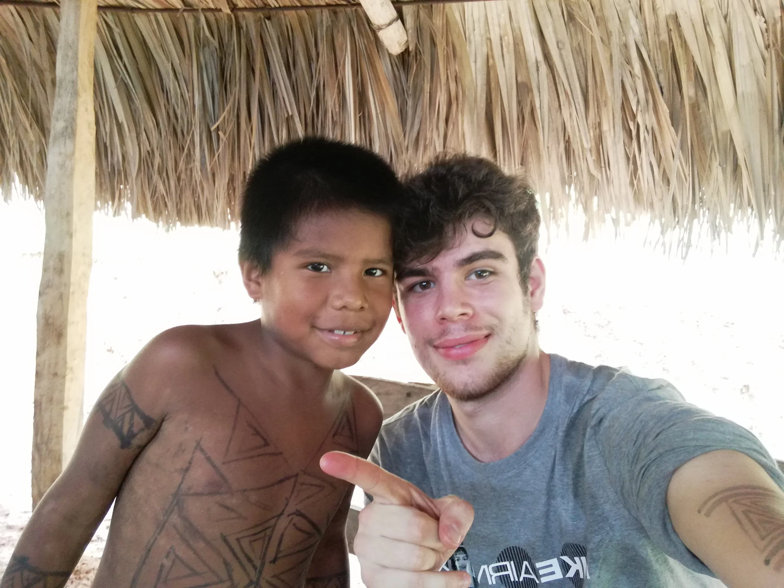 Jeune garçon autochtone et jeune homme caucasien posant ensemble sous un abri en chaume, souriants, dans un environnement en plein air.