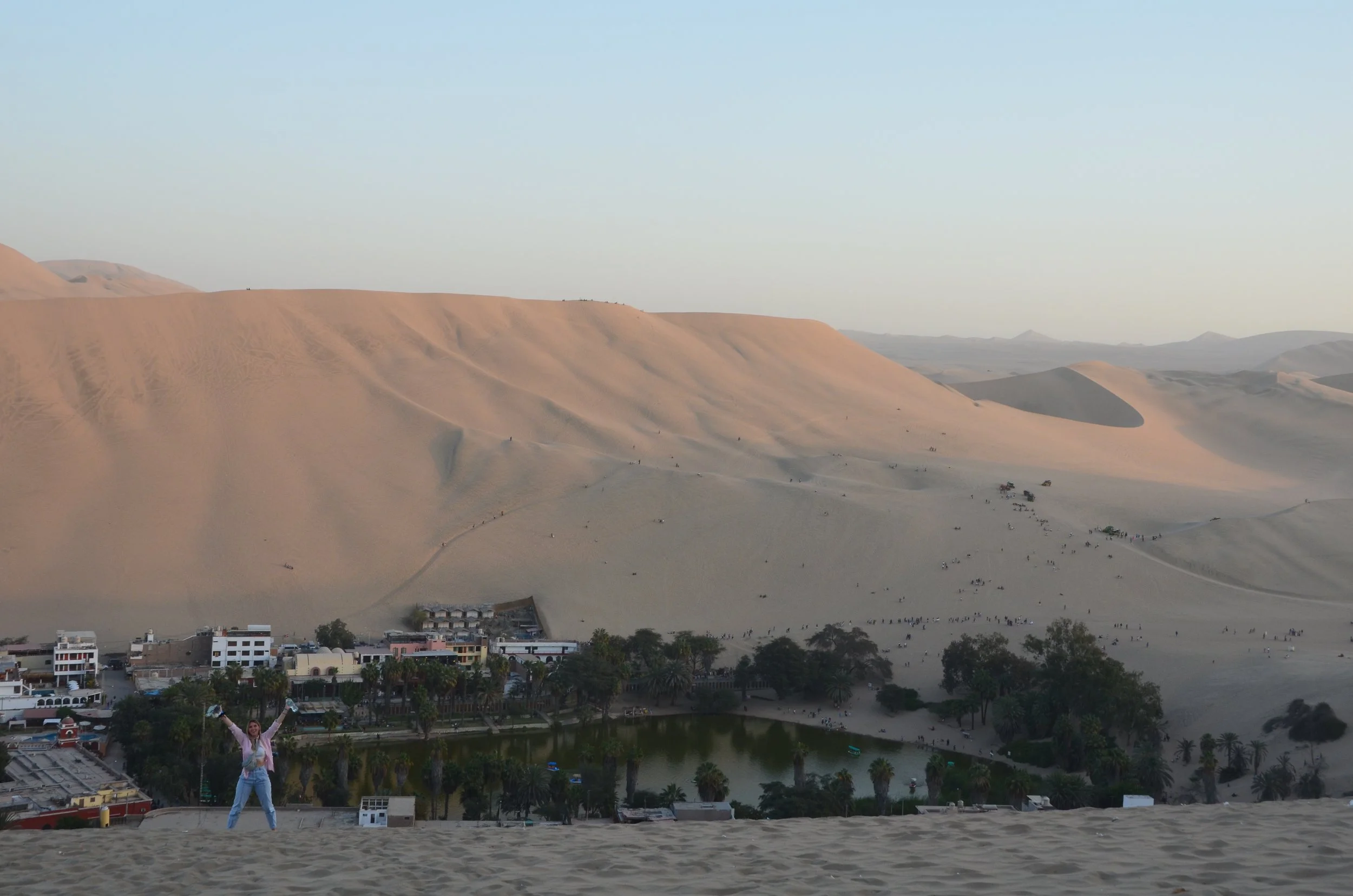 Paysage désertique avec dunes de sable, une ville en contrebas avec un lac, et une personne en premier plan avec les bras levés.