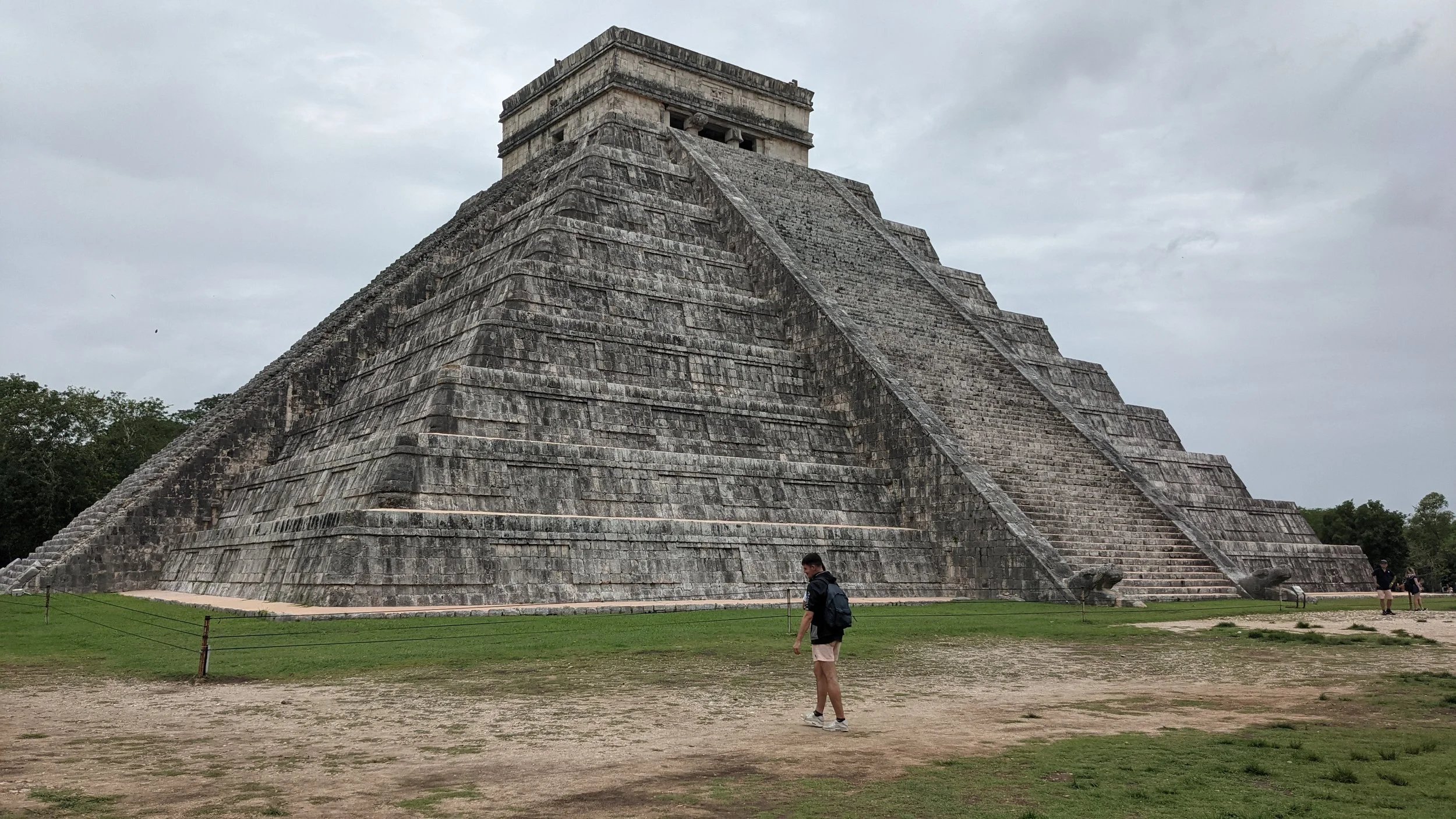 Une pyramide maya en pierre avec un escalier en pente sur un fond nuageux, entourée d'herbe et de quelques personnes.