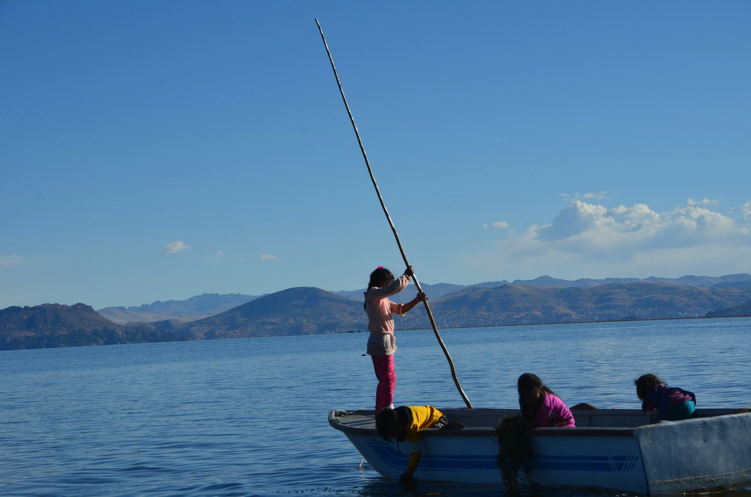 Enfants jouent dans un bateau sur un lac, avec des montagnes en arrière-plan, un ciel bleu et quelques nuages.